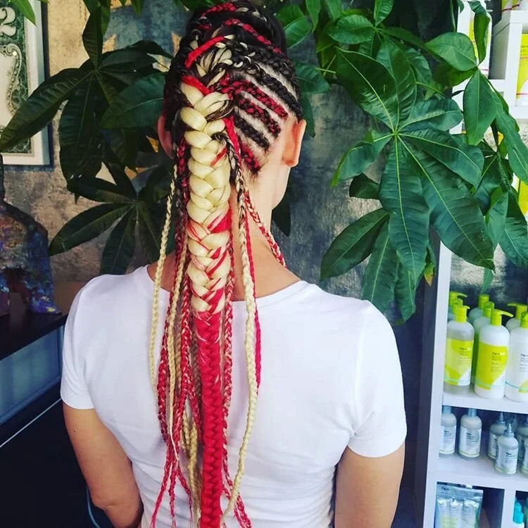 A woman with multi-colored braided hair standing in front of large green plants and a shelf with various bottles.