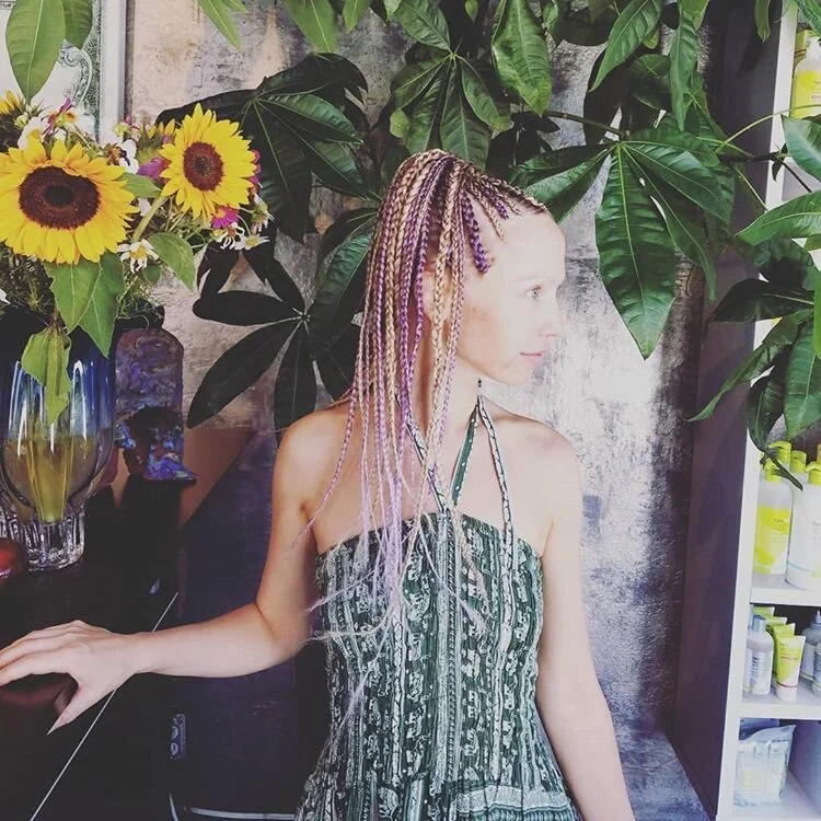 A woman with multicolored braided hair standing in front of tropical plants and a shelf with various bottles and containers, alongside a colorful sunflower arrangement in a glass vase.