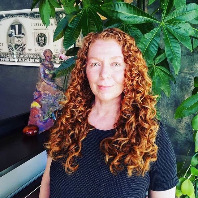 A woman with long, curly red hair posing indoors with lush green leafy plants behind her.