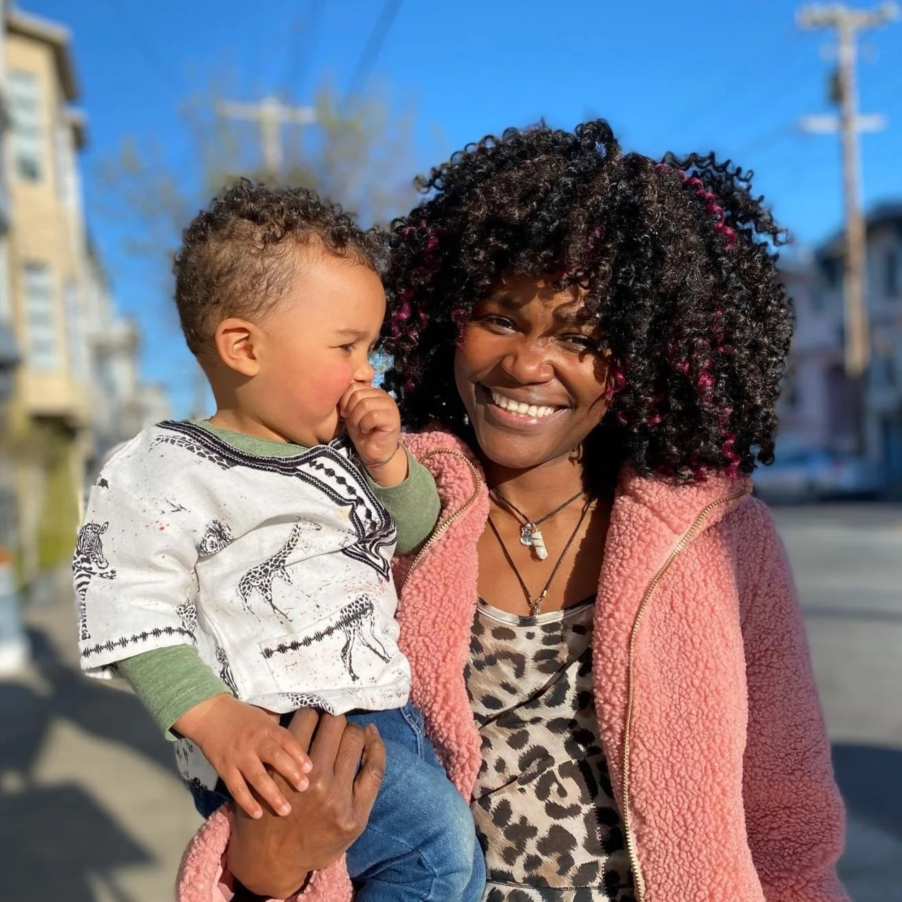 A smiling woman with curly hair holding a young child with short curly hair outdoors on a sunny day.
