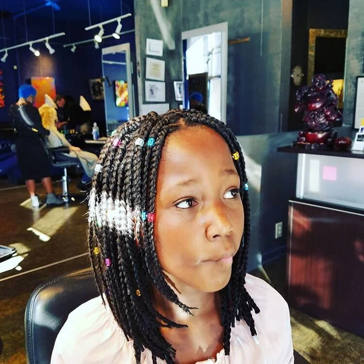 A young girl with braided hair and colorful beads, sitting in a hair salon, looking to the side with a neutral expression.