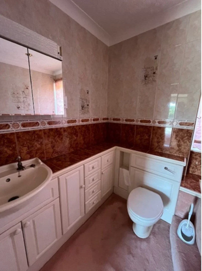 Original family bathroom with white fitted vanity cabinets, patterned tiles and pink carpet.