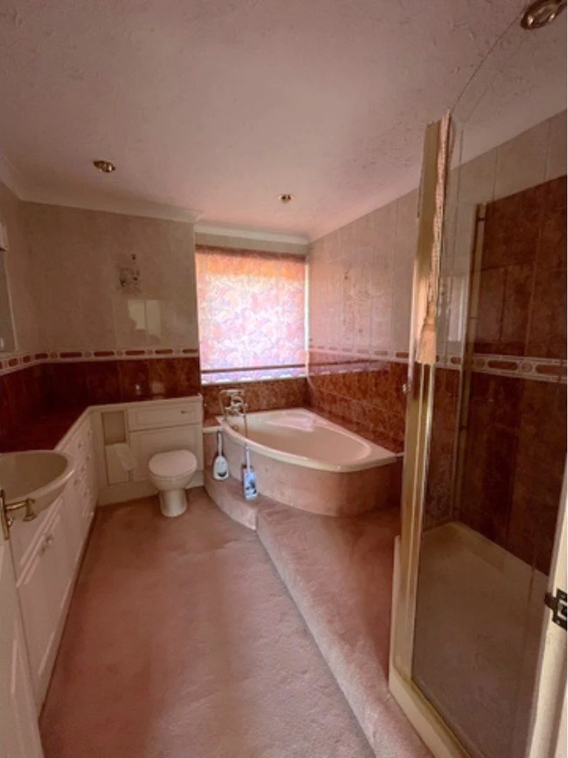 Family bathroom before renovation with raised corner bath, pink carpet and patterned wall tiles.