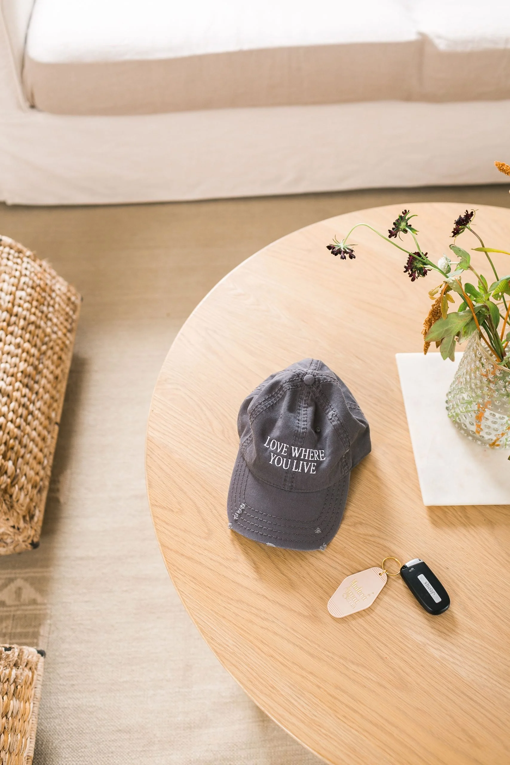 A round wooden coffee table with a black baseball cap that reads 'LOVE WHERE YOU LIVE', a set of car keys, and a glass vase with flowers. The background shows a beige sofa and a woven basket.