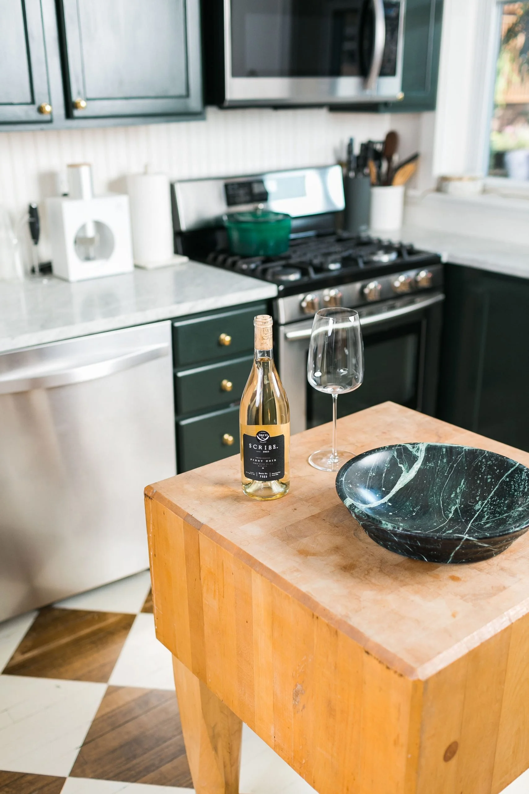 A kitchen with dark green cabinets, stainless steel appliances, and a wooden island. On the island, there is a bottle of white wine, a wine glass, and a decorative green marble bowl. The background shows a stove, microwave, and countertop with kitchen utensils.