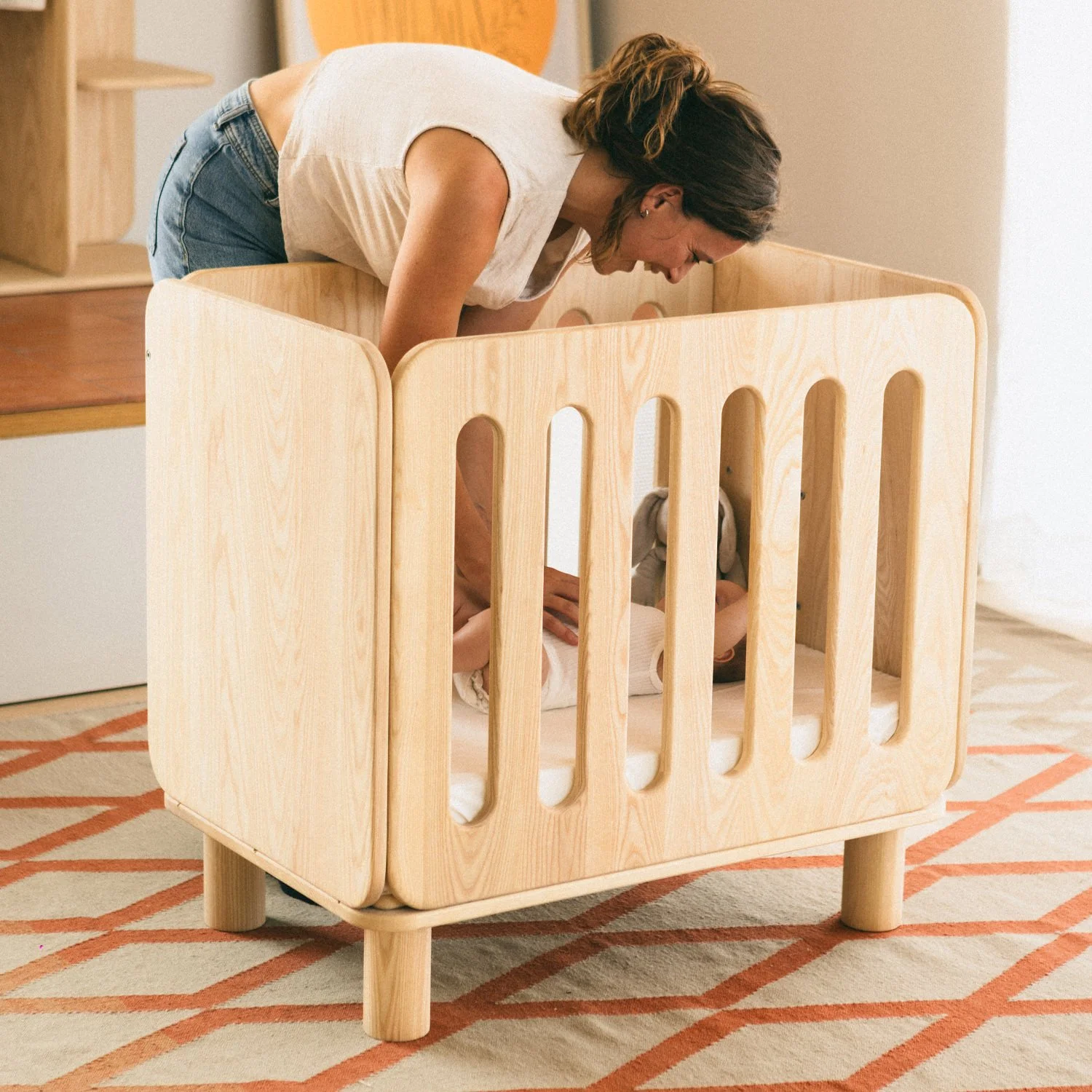 A mother interacting with a baby in the Nimbi bedside crib, highlighting the minimalist and serene design of the solid wood piece.