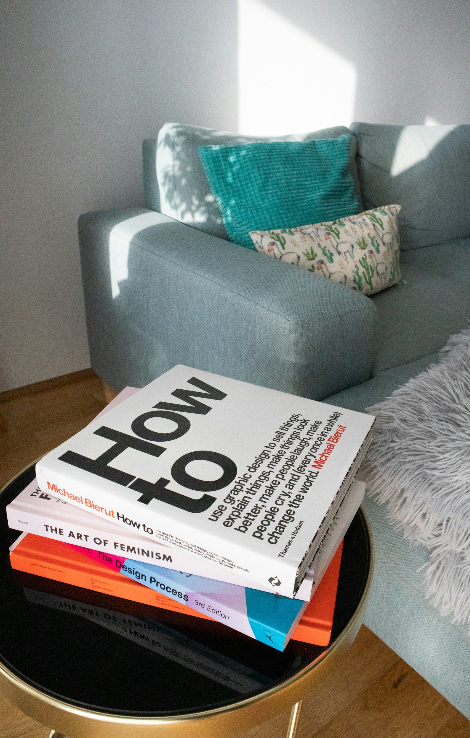 Stack of four books on a black and gold side table in front of a light blue sofa with patterned pillows, next to a gray shaggy blanket, in a living room with natural light coming through the window.