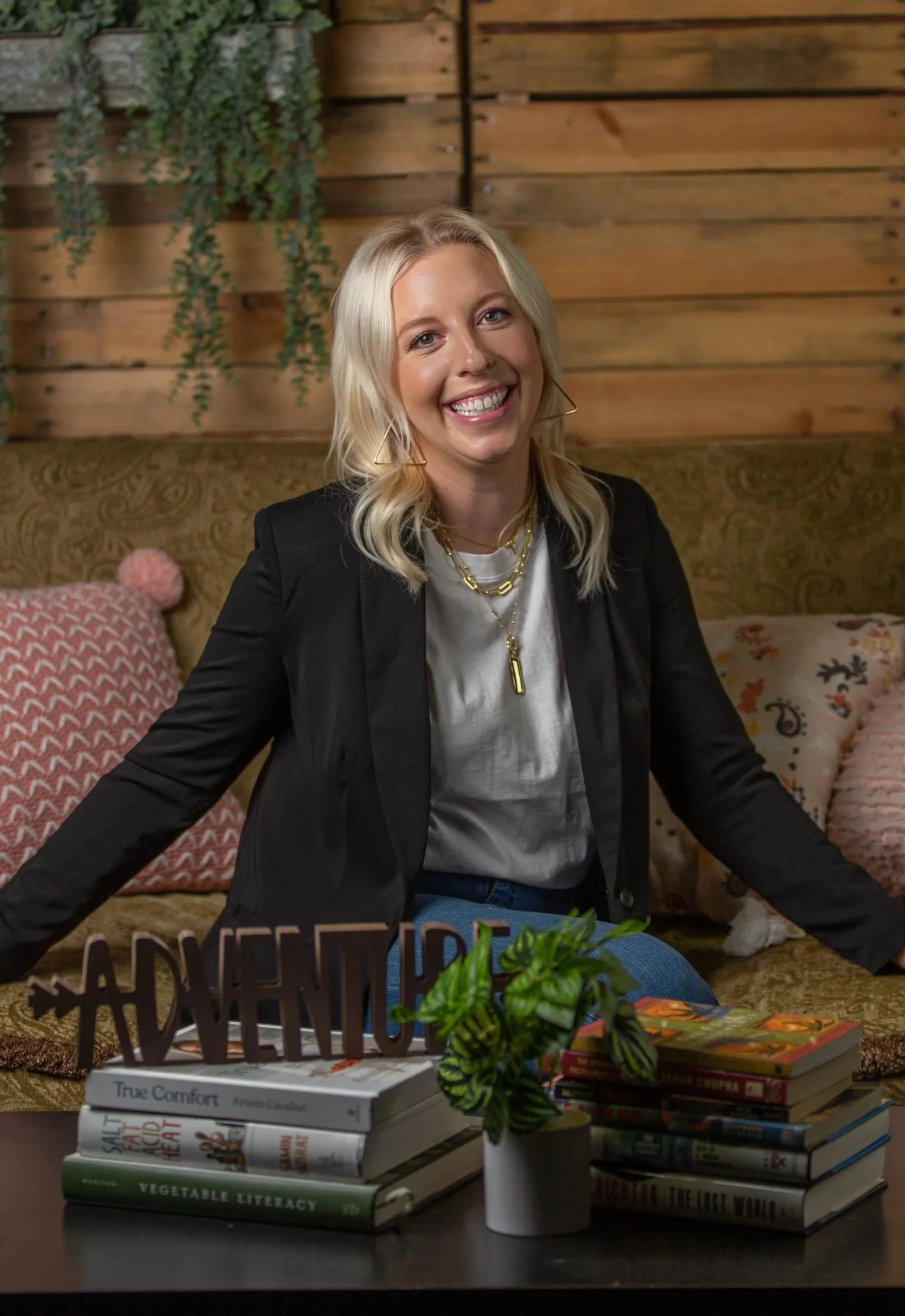 A smiling blonde woman in a black blazer sitting in a cozy room with a wooden wall background, surrounded by pillows and books on a table in front of her.