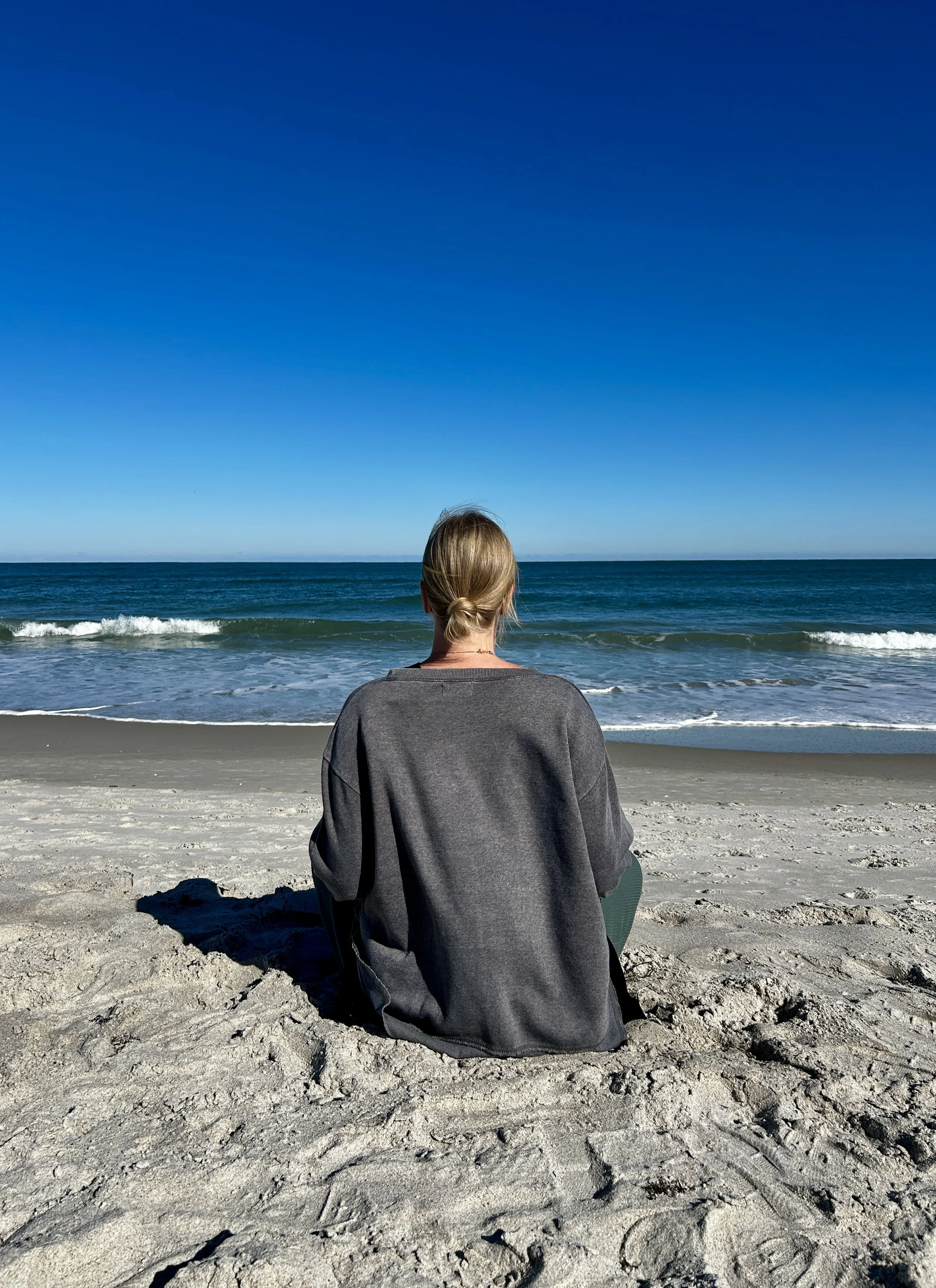 Person sitting on the sandy beach facing the ocean, with blue sky above and waves in the background.