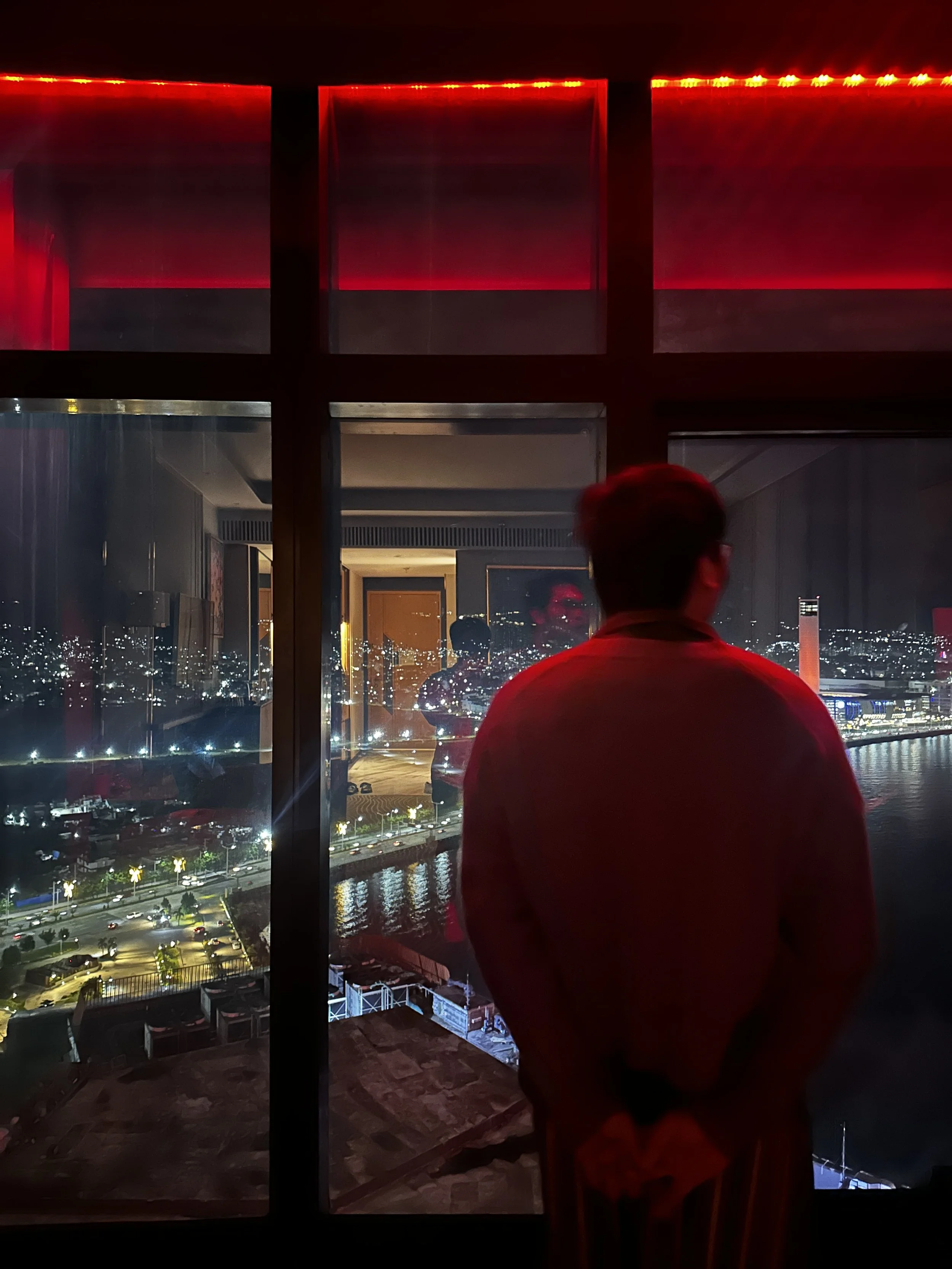 Person with glasses looking out a large window at a city nightscape with lights reflecting on the water.