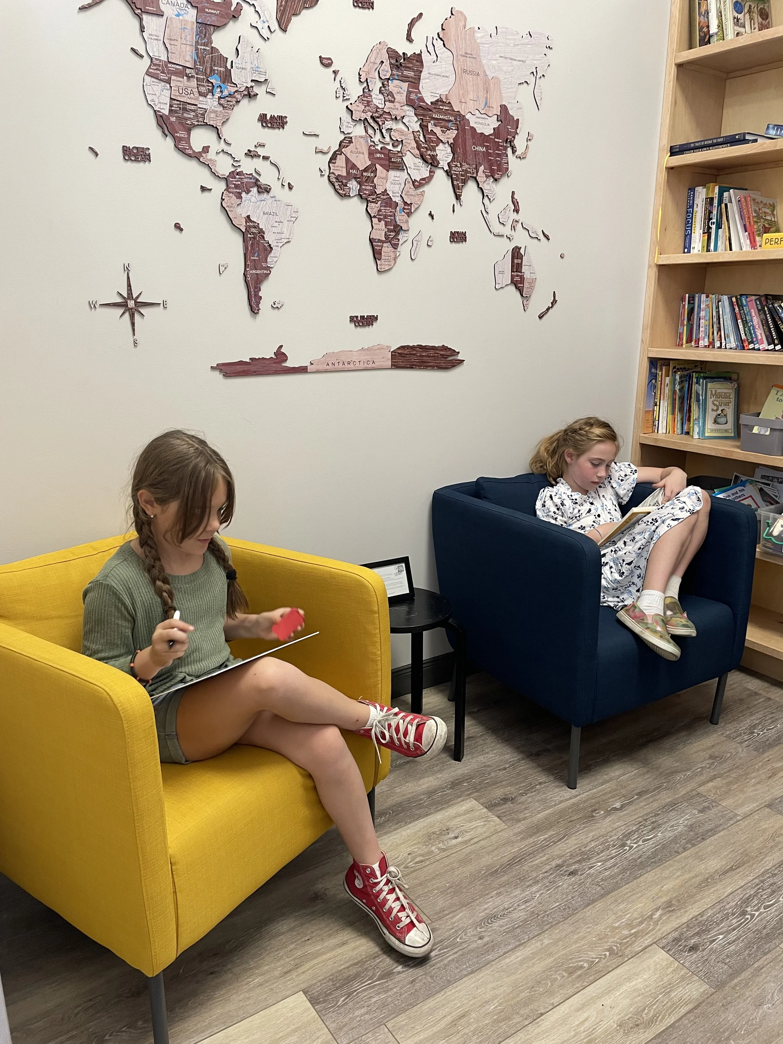Two young girls sitting on colorful armchairs in a cozy room, reading and drawing. A world map is on the wall behind them, and a bookshelf filled with books is to the right.
