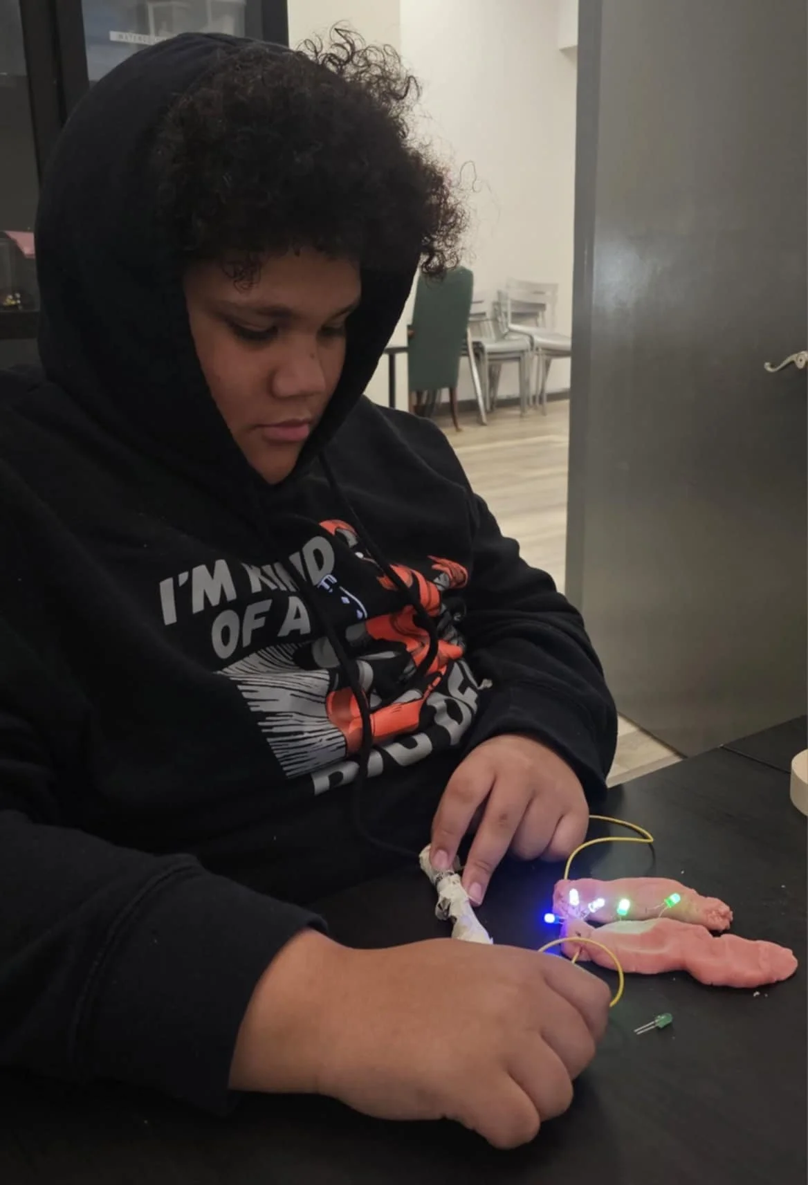 A young person sitting at a table with a panda-shaped plush toy modified with LED lights and electronic components, practicing or demonstrating a hands-on project.