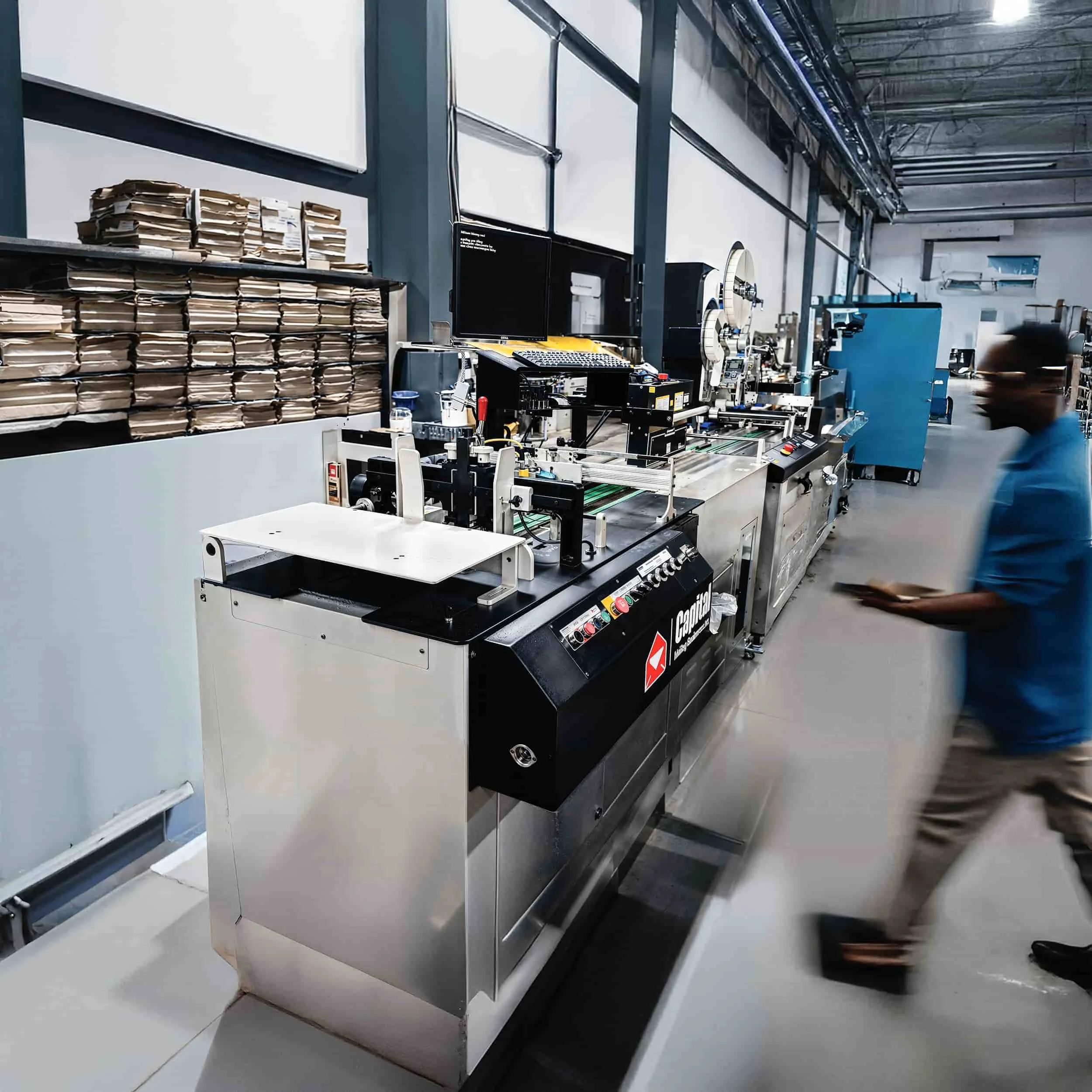 Industrial manufacturing facility with machinery, electronic controls, and stacks of paper or flat materials. A person in a blue shirt and beige pants is walking past the equipment.