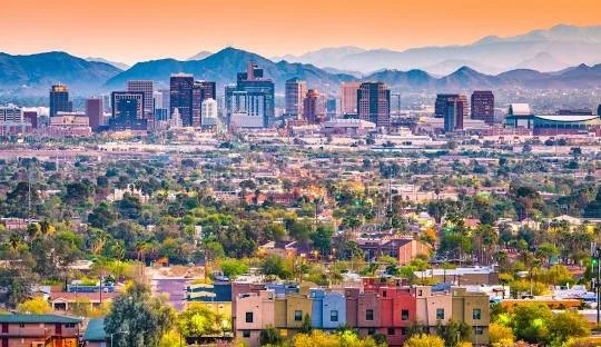 Skyline of Phoenix, Arizona, with downtown buildings, green vegetation, and distant mountains at sunset.