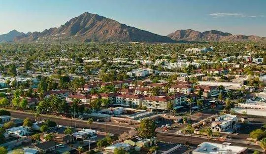 Aerial view of a cityscape with numerous buildings, streets, and green spaces, with rugged mountains in the background.