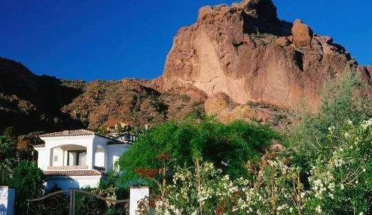 A large rocky mountain with a white house and green trees and shrubs in the foreground under a clear blue sky.