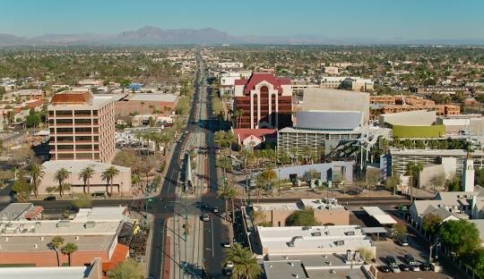 Aerial view of a cityscape with a main road flanked by commercial buildings, palm trees, and distant mountains in the background.