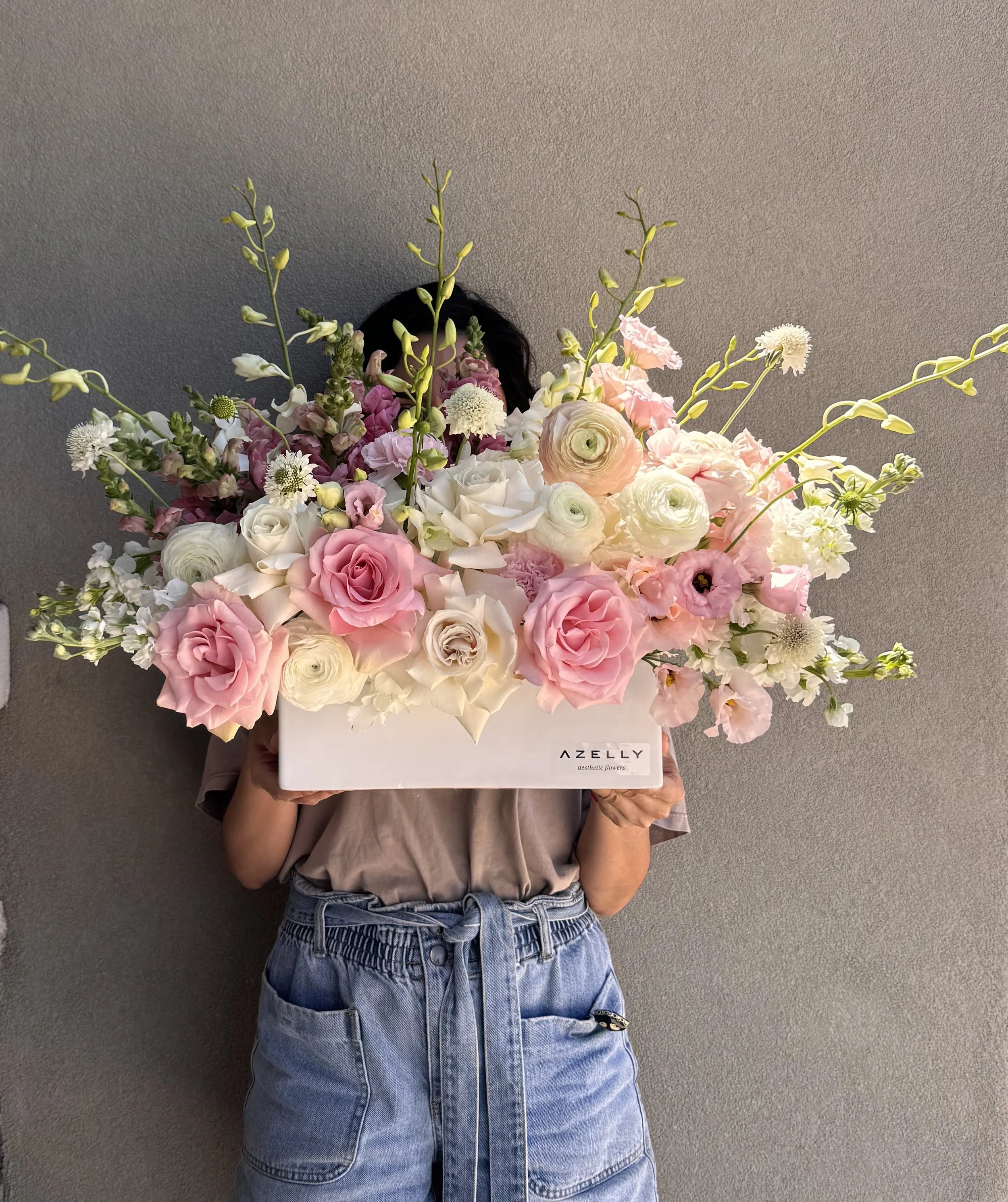 Person holding a large bouquet of pink, white, and yellow flowers in front of their face, wearing a gray shirt and high-waisted blue jeans, against a gray wall.