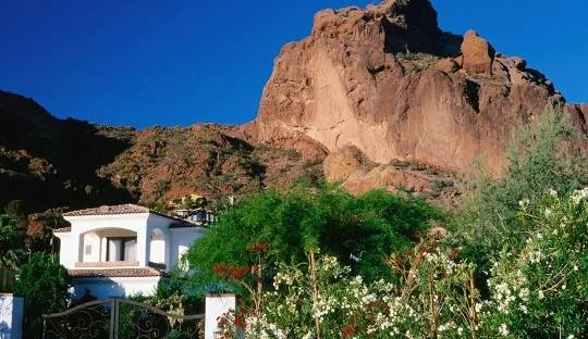White house with a tile roof near a rocky mountain and lush greenery under a clear blue sky.