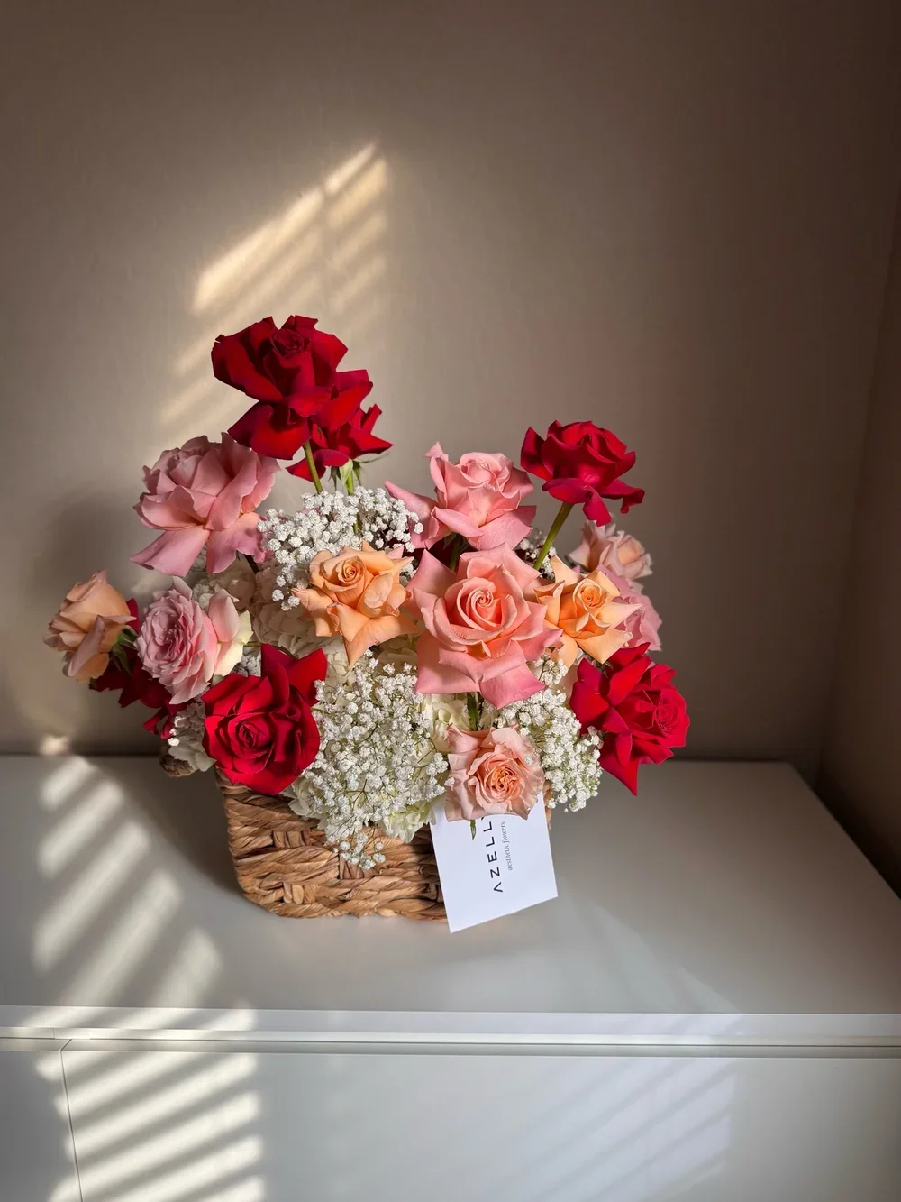 A basket filled with pink, red, and peach roses, along with white baby's breath flowers, placed on a white surface against a wall with sunlight streaming through blinds.