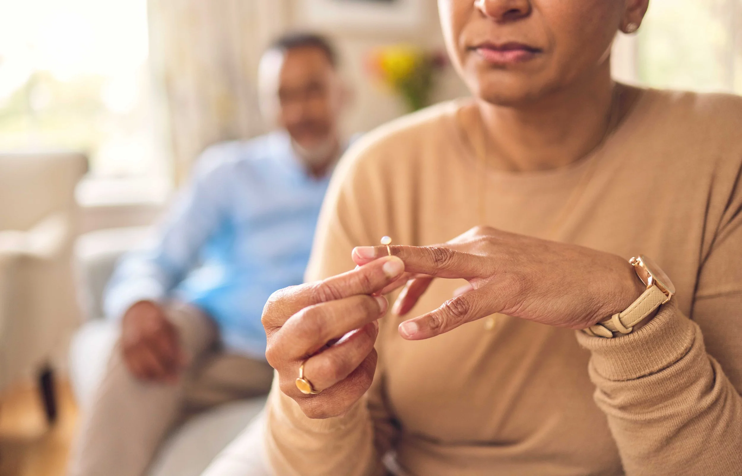 Older woman removing her wedding ring while a man sits out of focus in the background, suggesting a couple facing separation