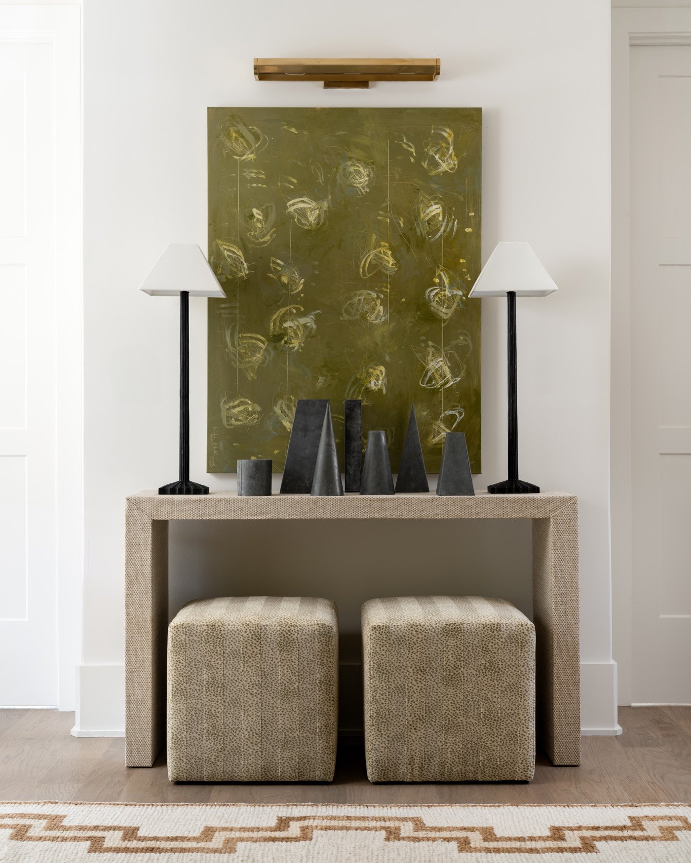 Interior scene with a neutral grasscloth console table, two animal print ottomans, black fluted lamps, zinc geometric sculptures, a large green abstract painting, and a patterned rug.