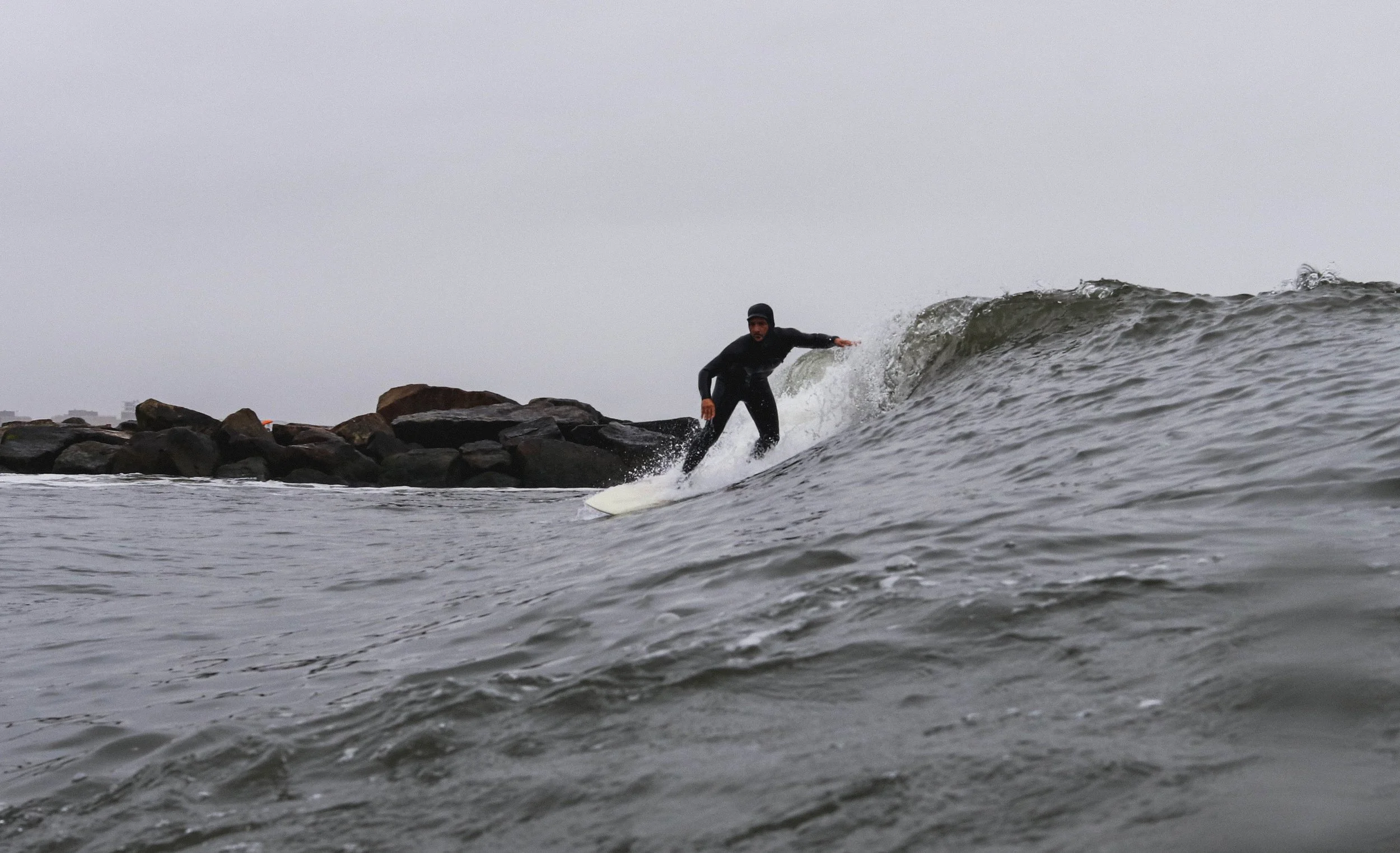 A person wearing a black wetsuit and black beanie surfing on a small wave near rocks on a cloudy day.