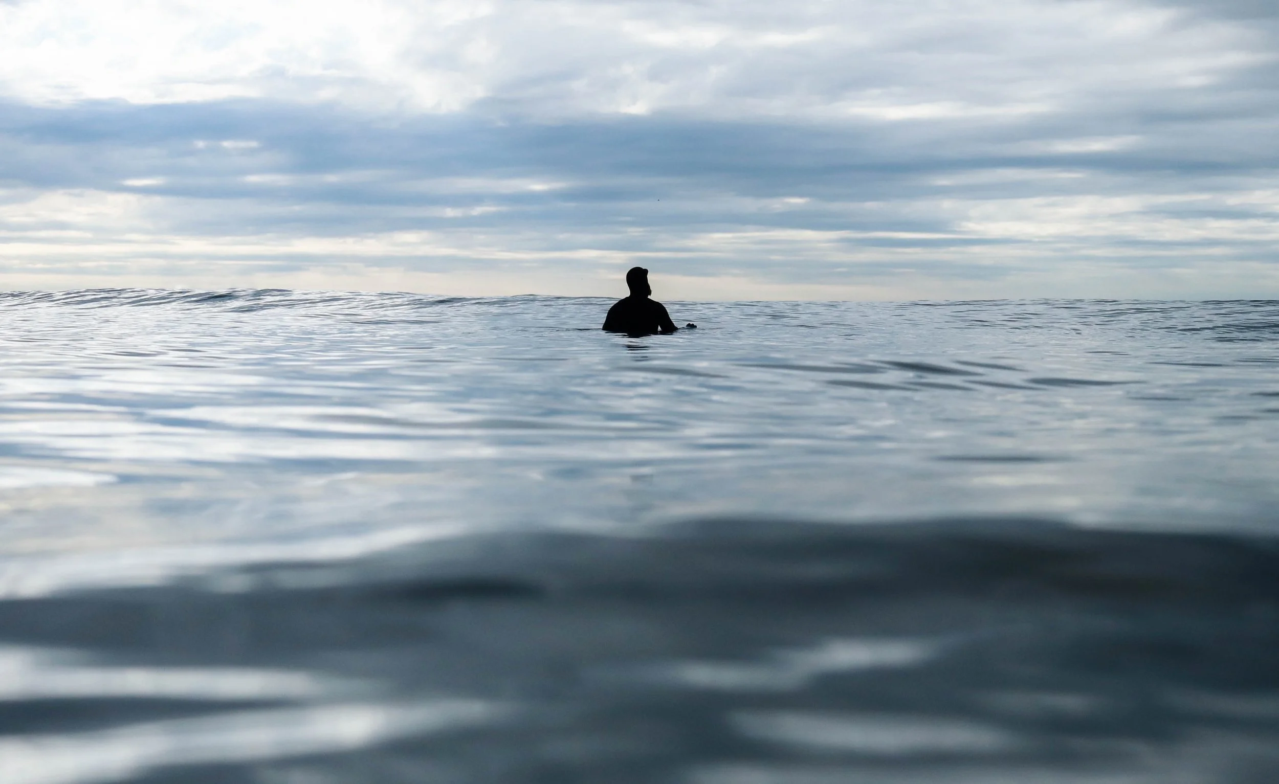 A person with short hair sitting in calm ocean water, facing the horizon under cloudy sky.