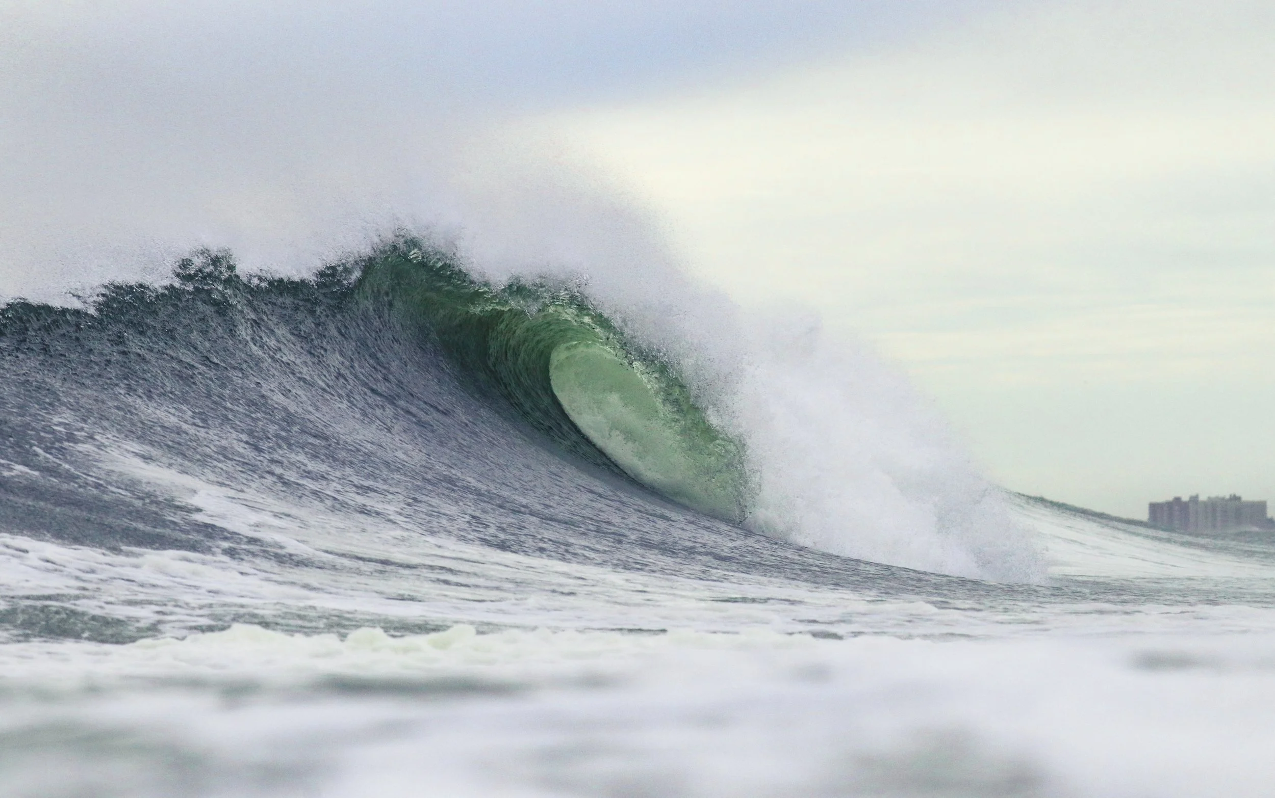 A large ocean wave in the process of breaking, with high surf and spray, near a distant shoreline with some buildings in the background.