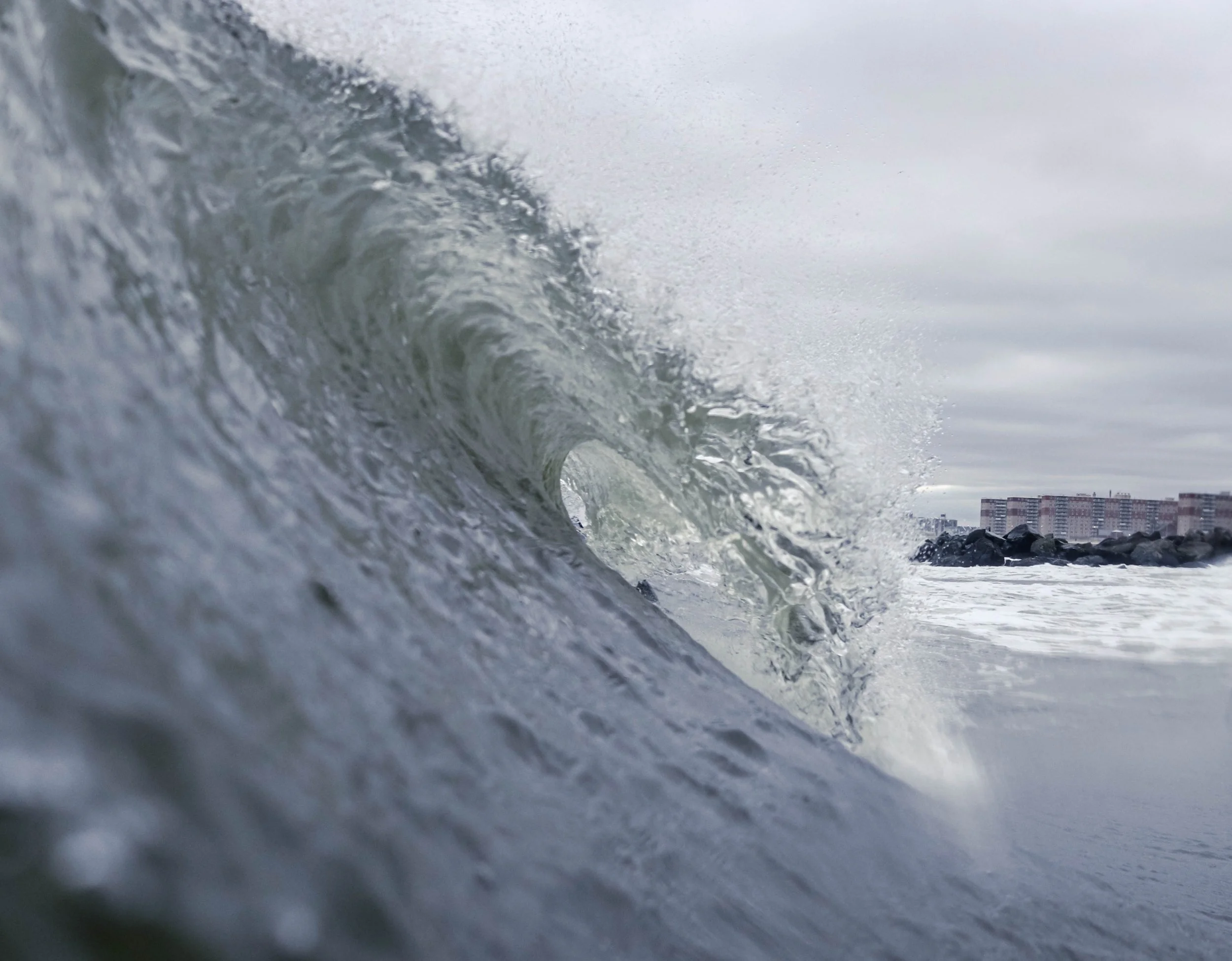 A large ocean wave crashing with buildings visible in the distance under an overcast sky.
