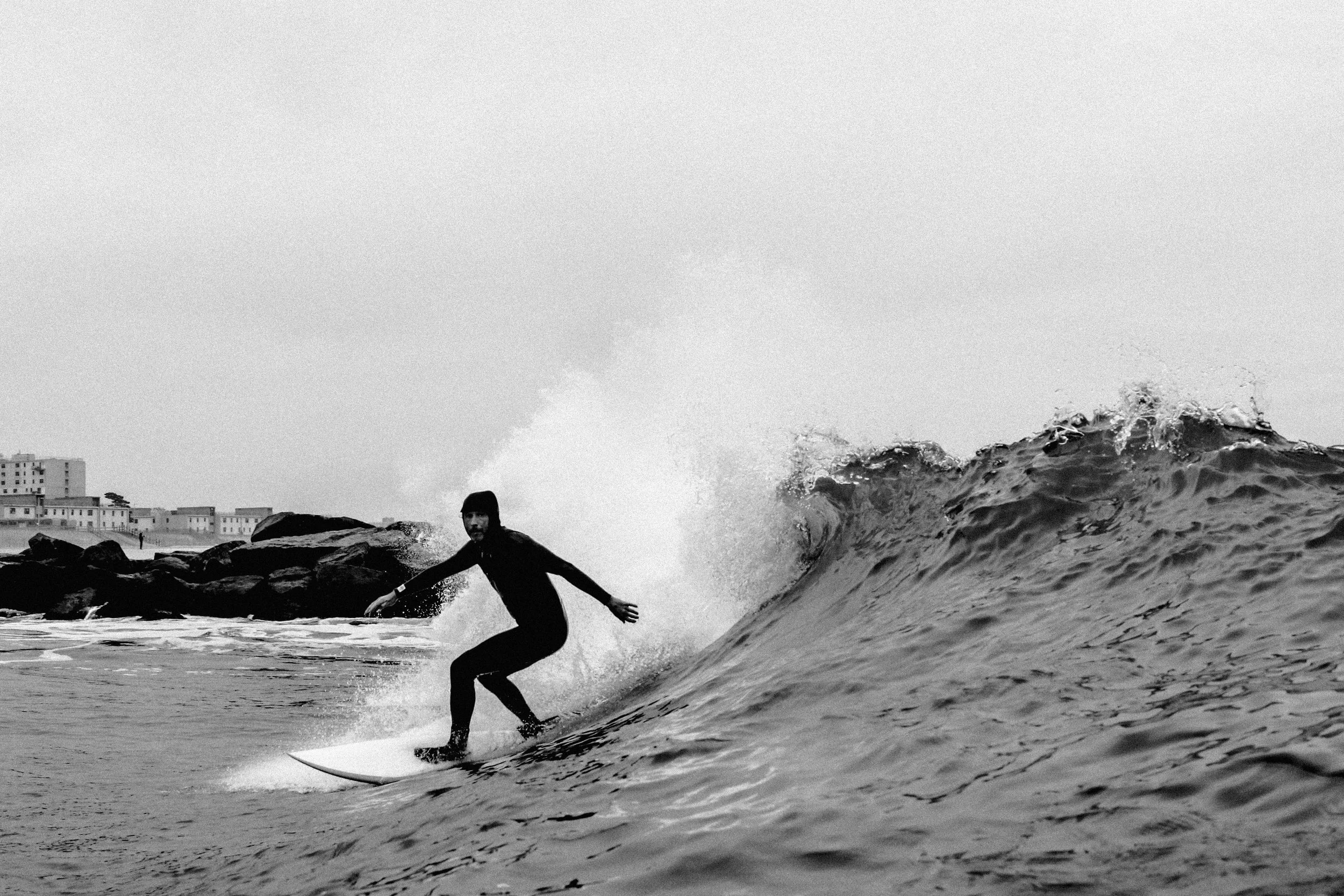 A person surfing on a wave at the beach, with buildings visible in the background, in black and white.