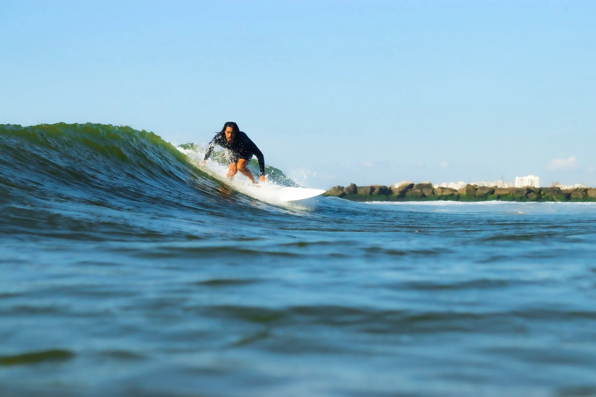 A person surfing on a wave in the ocean during daytime with clear sky, rocks, and buildings in the background.