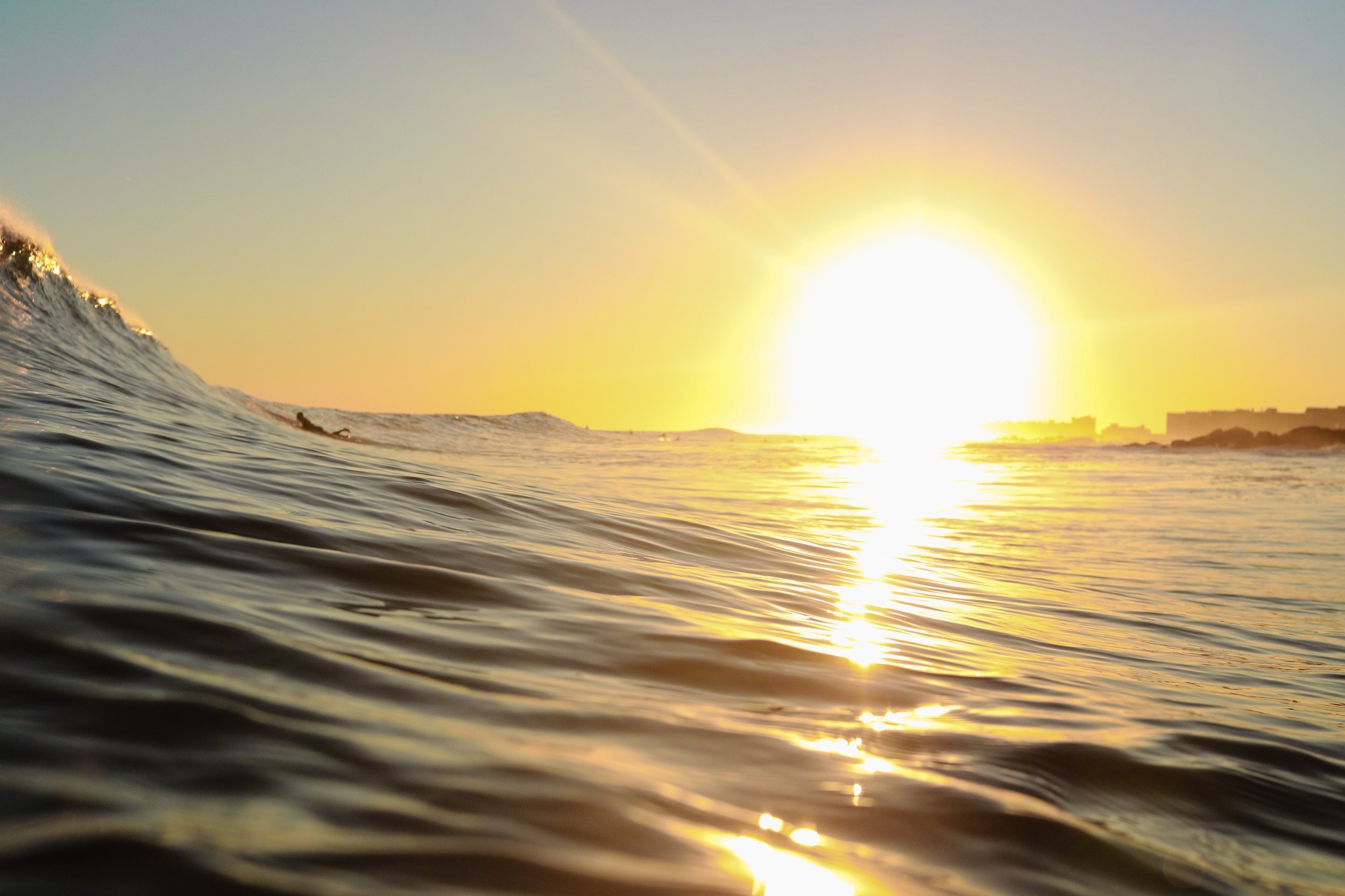 Sunset over ocean with water ripples and a silhouetted surfer in the distance.