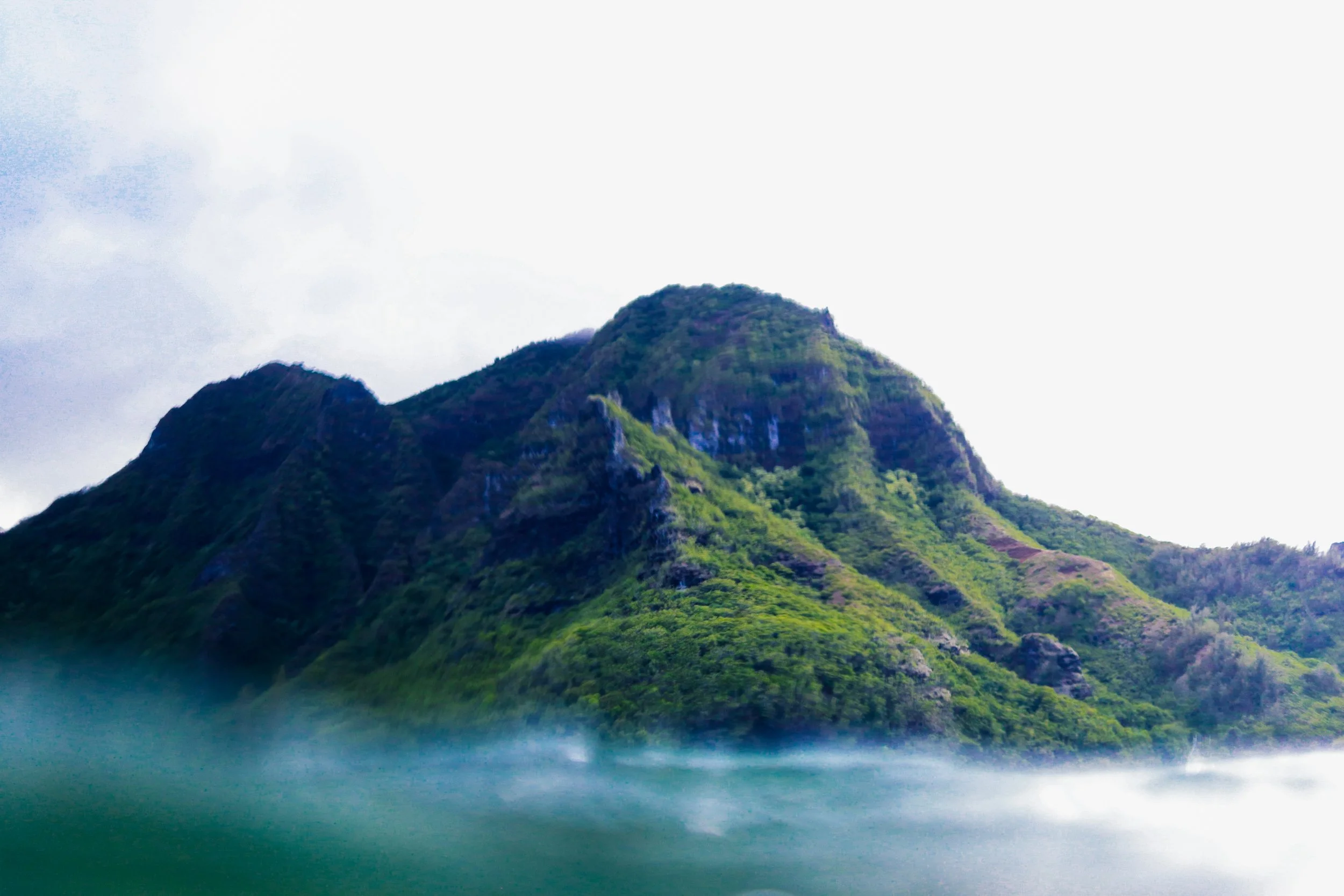 A green mountain with lush vegetation rising from a body of water under a cloudy sky.