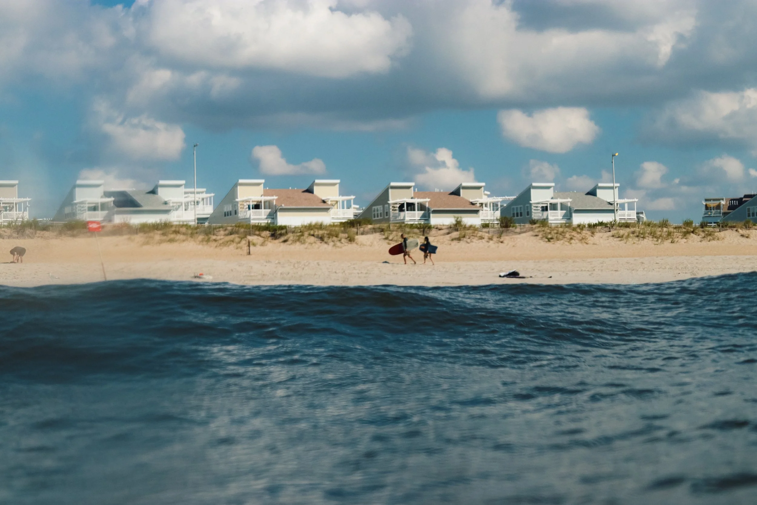 View of a sandy beach with several white beach houses and two surfers carrying surfboards, under a partly cloudy sky.