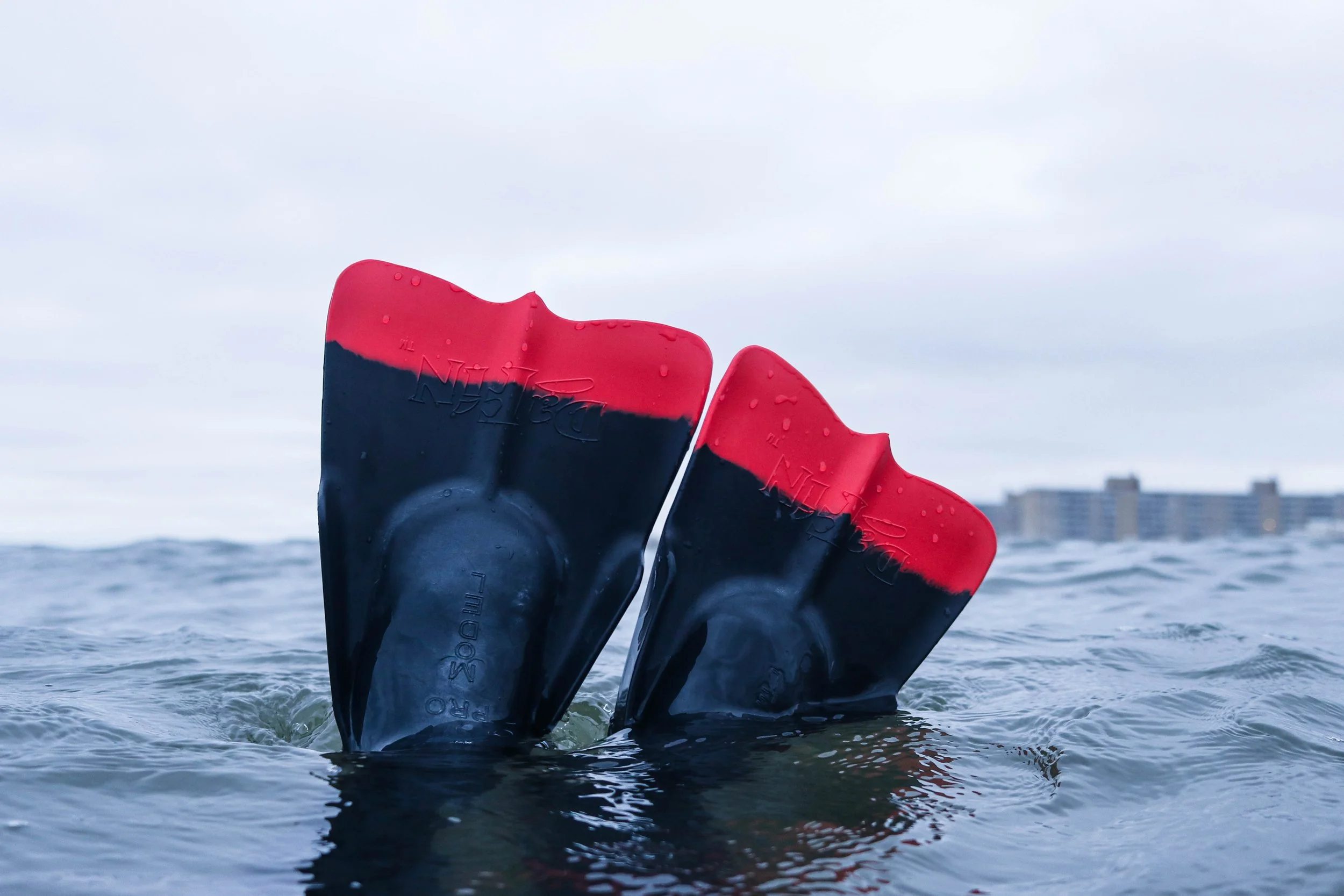 Close-up of black and red fins of a swimmer emerging from the water during a race, with a cloudy sky and city buildings in the background.