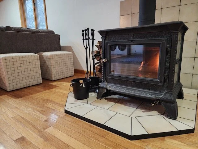 A black wood stove with a glass door showing a fire inside, positioned on a tiled hearth in a living room, with fireplace tools and a bucket nearby, and a beige couch with plaid ottomans in the background.