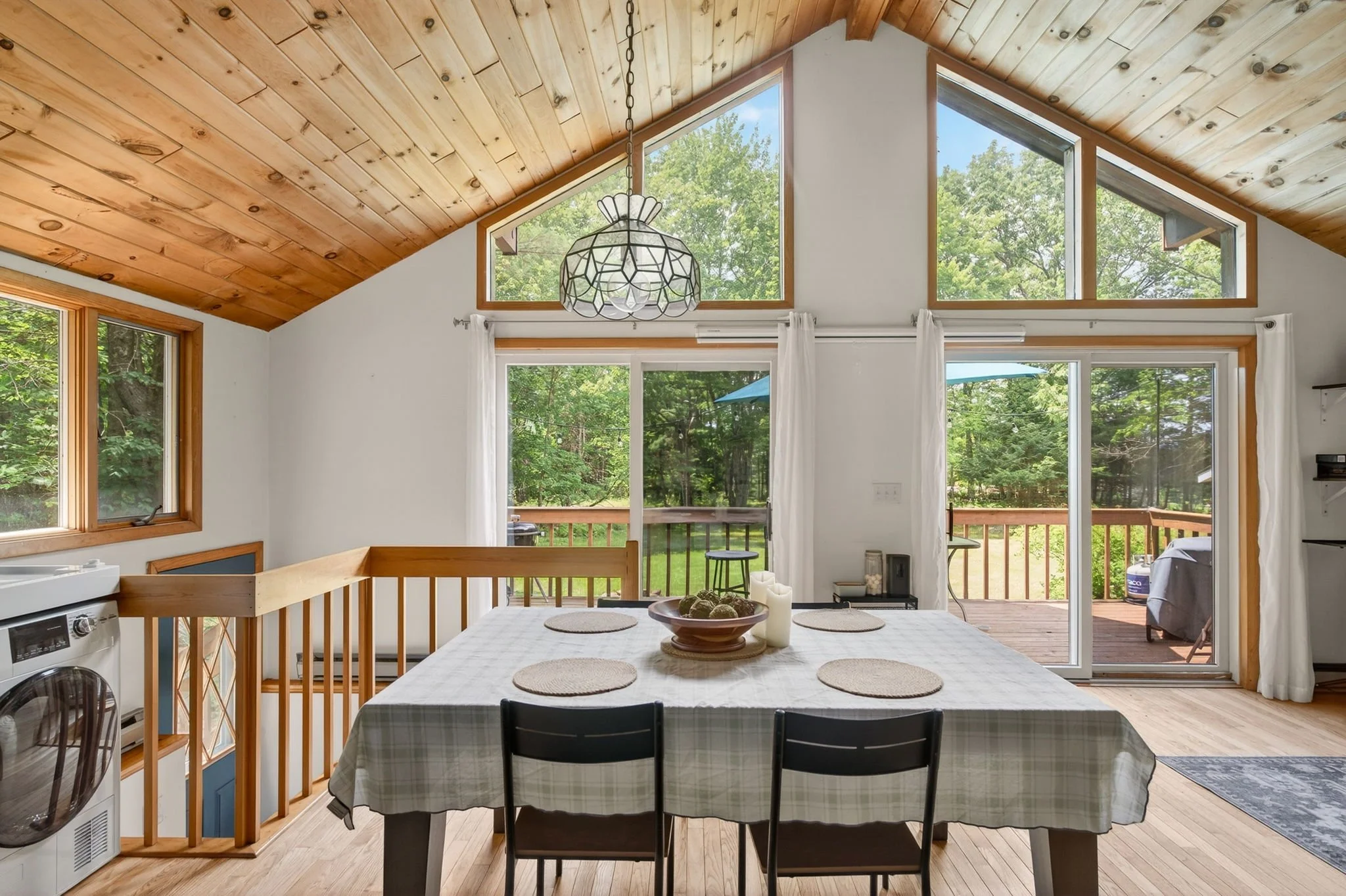 Dining area in a house with large windows and sliding glass door leading to a deck. The room has a wooden ceiling, white walls, and a table with a checkered tablecloth and four placemats. Outside the deck, there is a grill and some outdoor furniture, and the scene is surrounded by green trees.