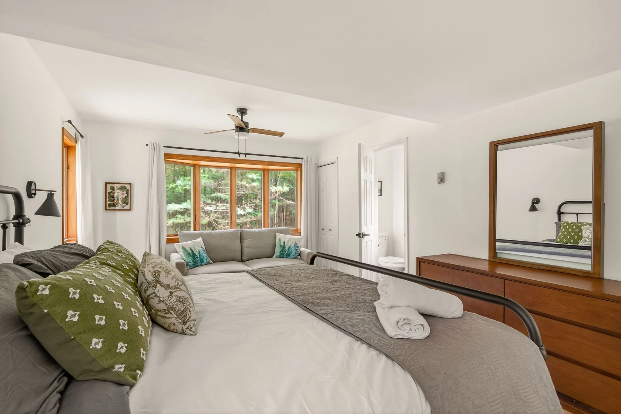 A cozy bedroom with a large bed, decorated with green, beige, and patterned pillows, a wooden dresser with a mirror, a beige sofa with teal pillows near a window, and a ceiling fan. The room has white walls and wooden window frames, overlooking a wooded outdoor area.