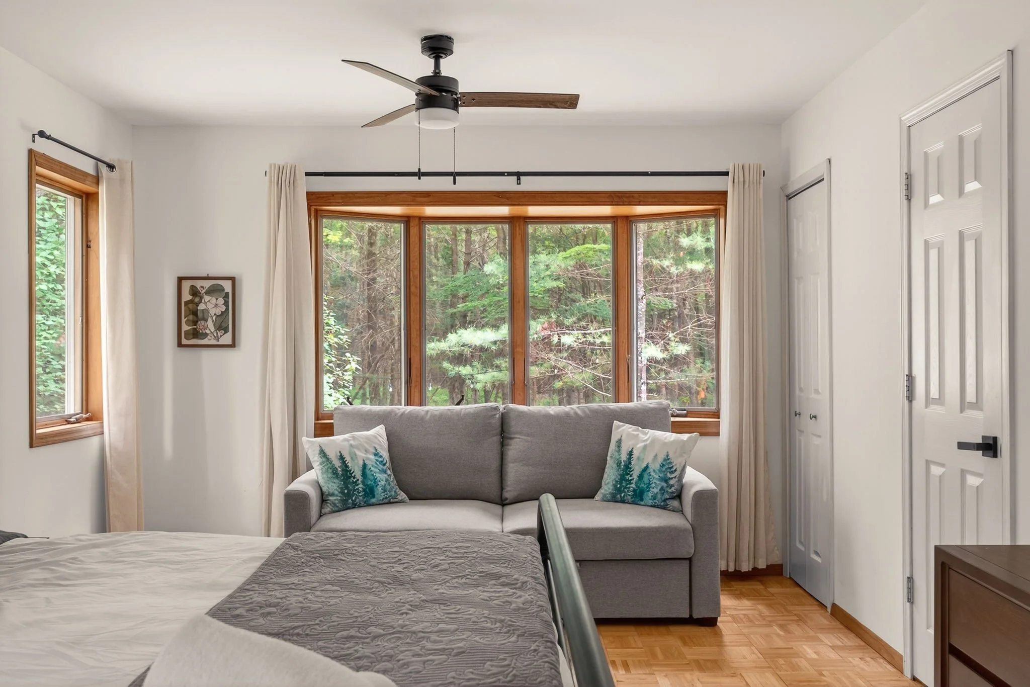 A bedroom with a bed, a gray sofa with decorative pillows, and windows showing a wooded outdoor view. There is a ceiling fan and wooden trim around the windows and a closet door.