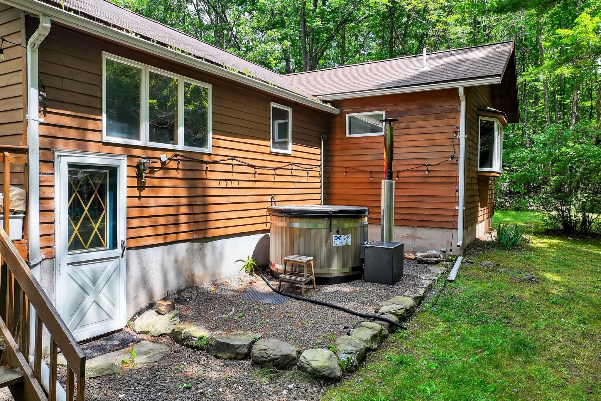 Backyard of a house with wooden siding, a hot tub, string lights, and surrounding trees.