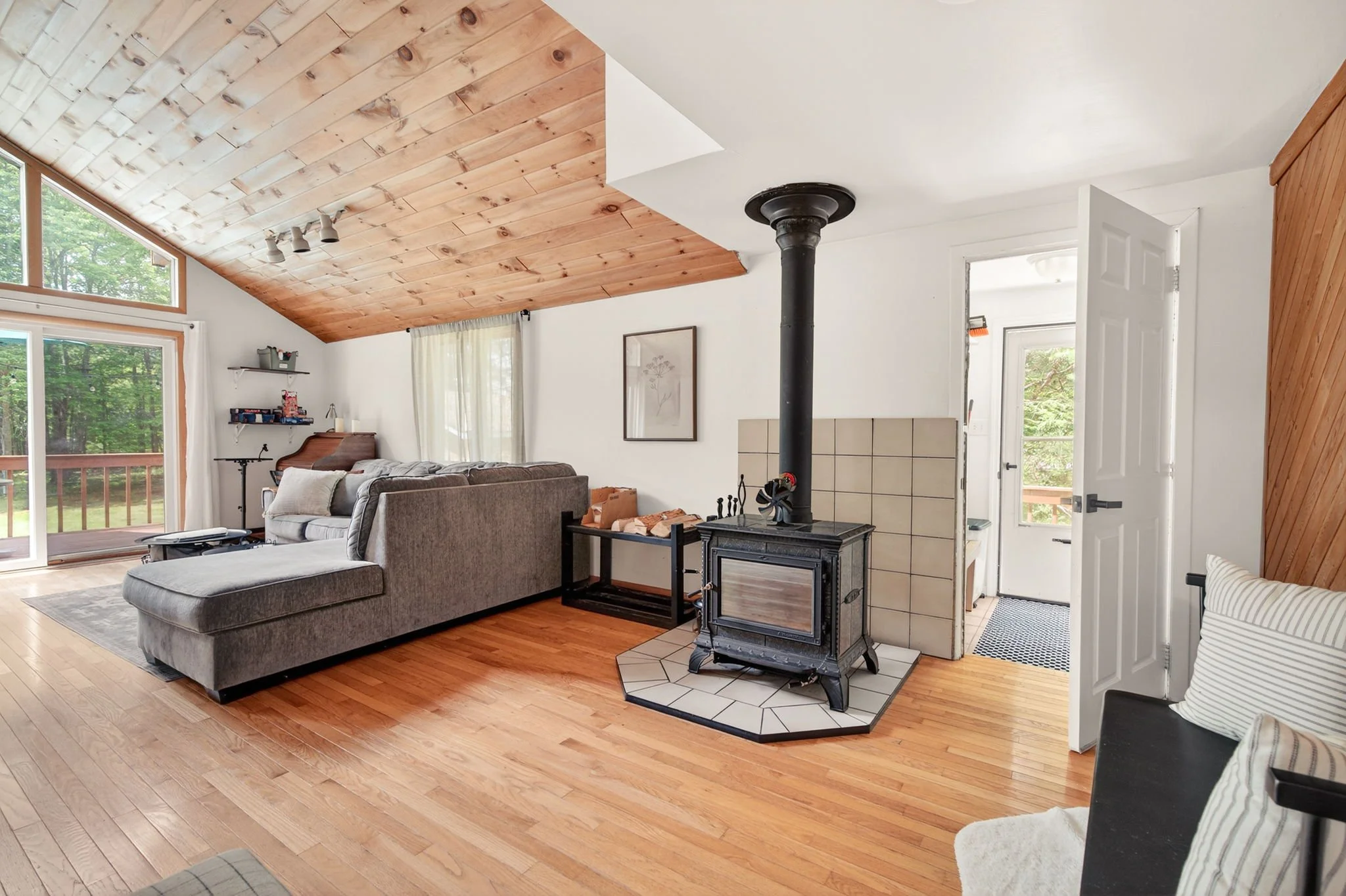 Living room with a gray upholstered sectional sofa, large sliding glass door leading to a wooden deck, wood-paneled ceiling, white walls, a framed picture on the wall, a black wood stove with a stovepipe, and a doorway to a sunlit outdoor area.