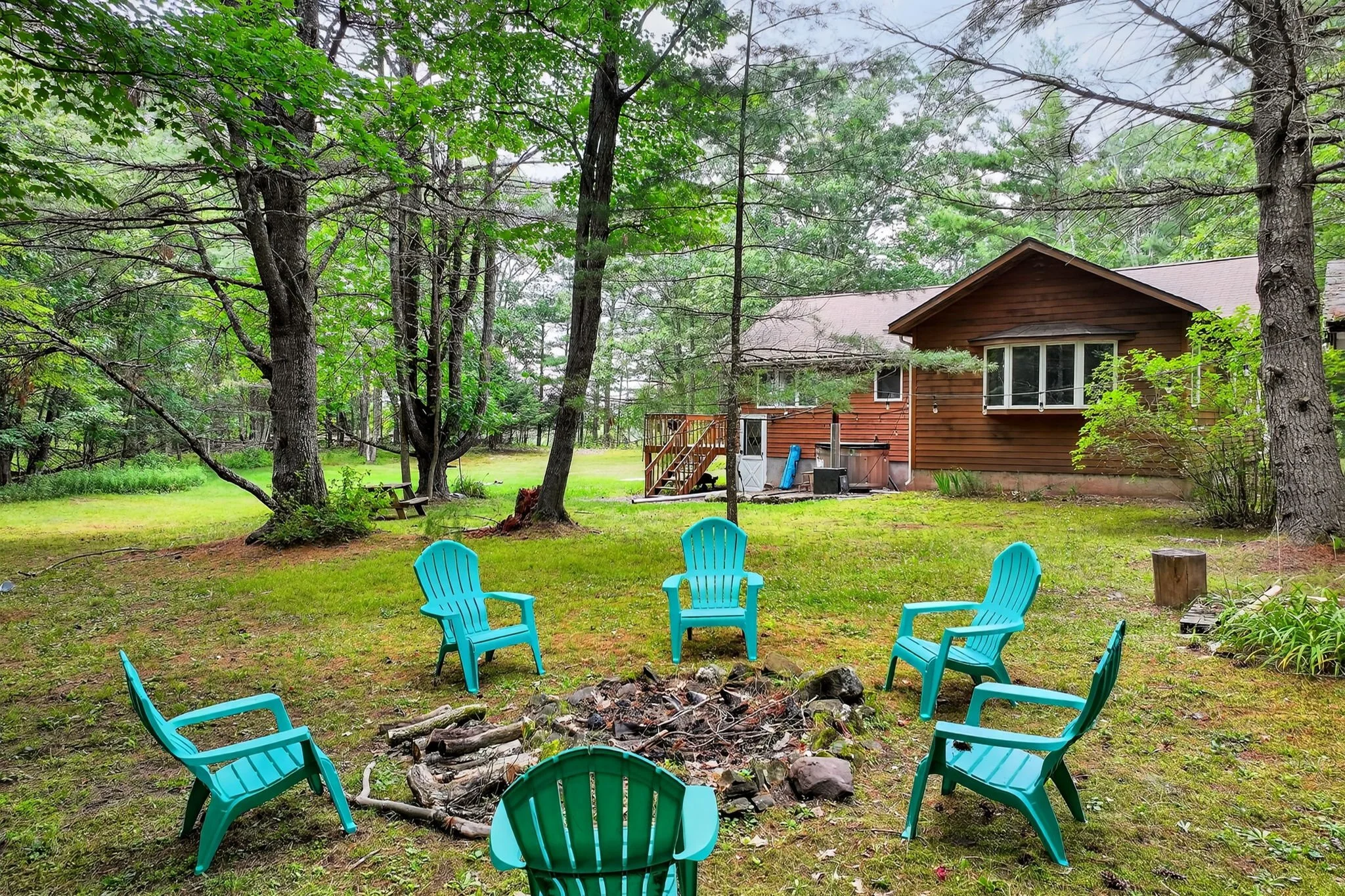 Backyard with six blue Adirondack chairs arranged in a circle around a fire pit with logs. A grassy yard with trees and a brown house in the background.