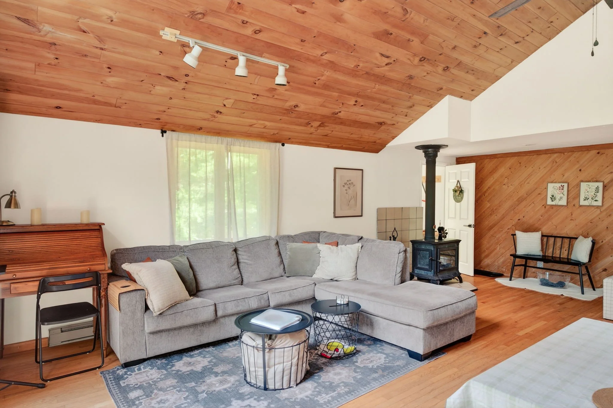 Living room with a gray sectional sofa, wooden ceiling, and white walls. A black wood stove, chair, and decorated wooden wall panels are visible.