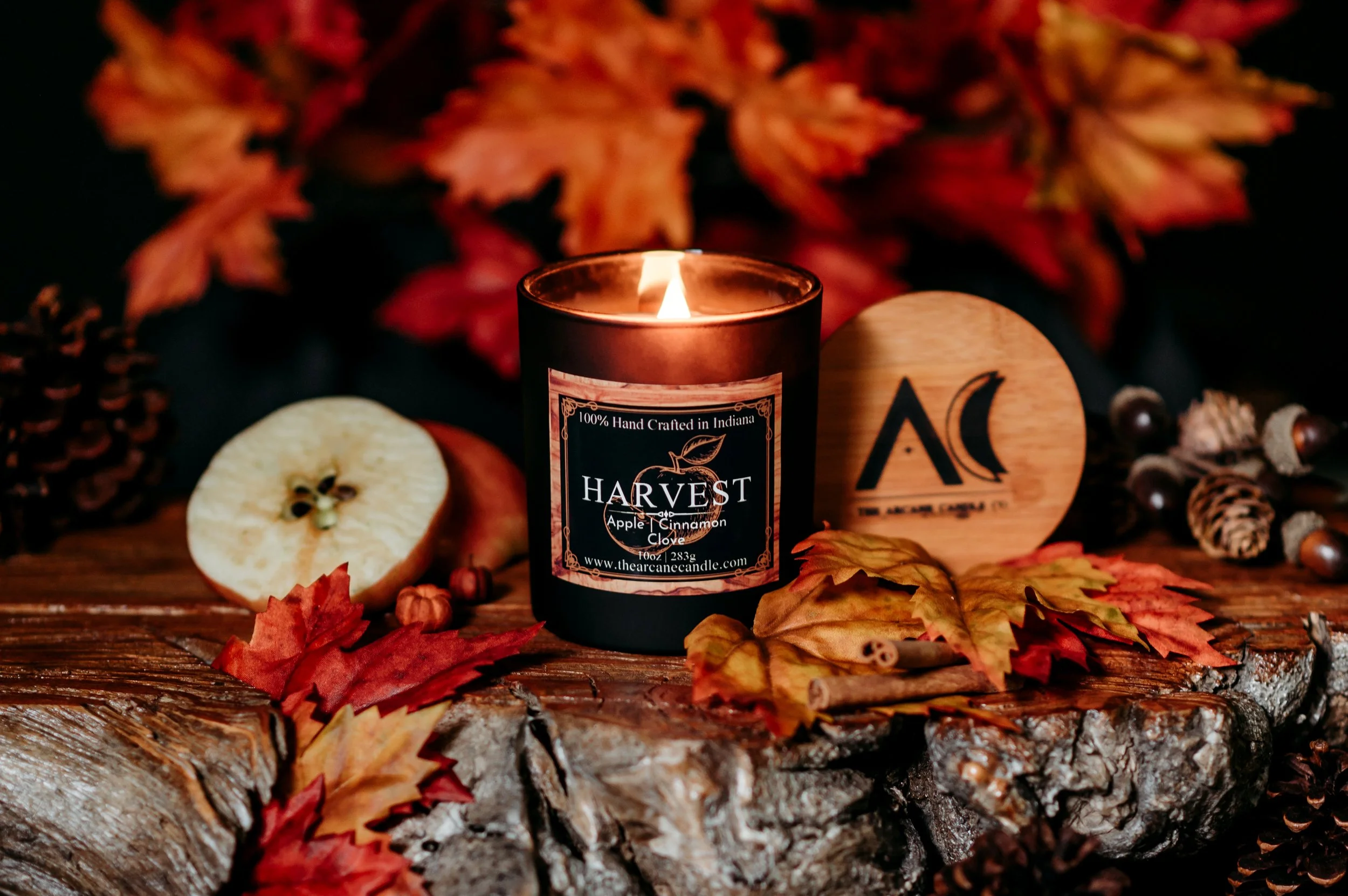 HARVEST baked apple candle in a matte black jar styled with fresh apple, cinnamon sticks and fall foliage.