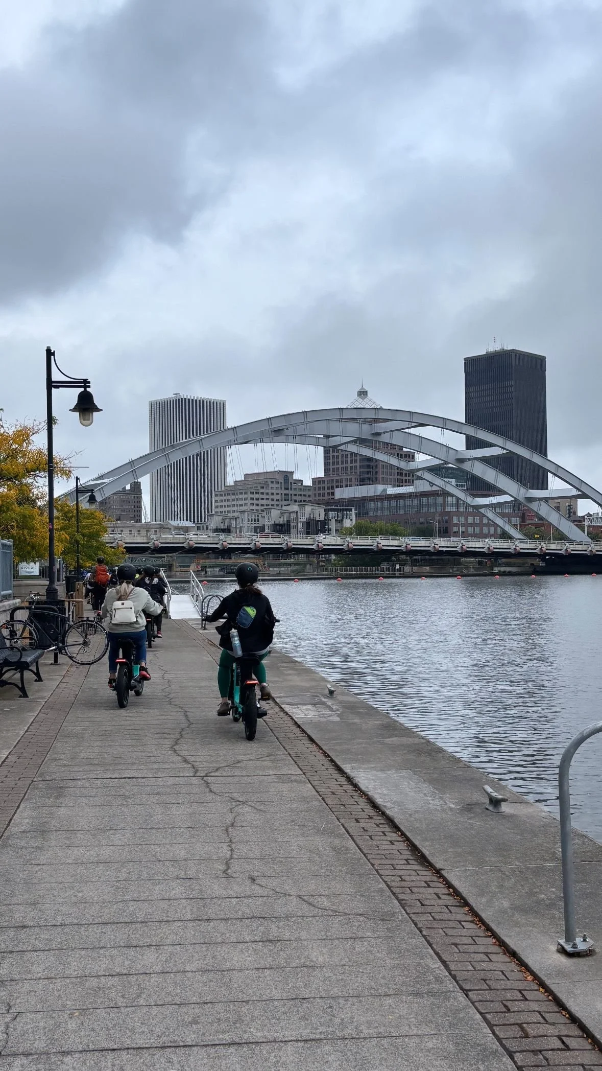 alt="a group of bicyclists from behind bike along a riverfront on a cloudy day."