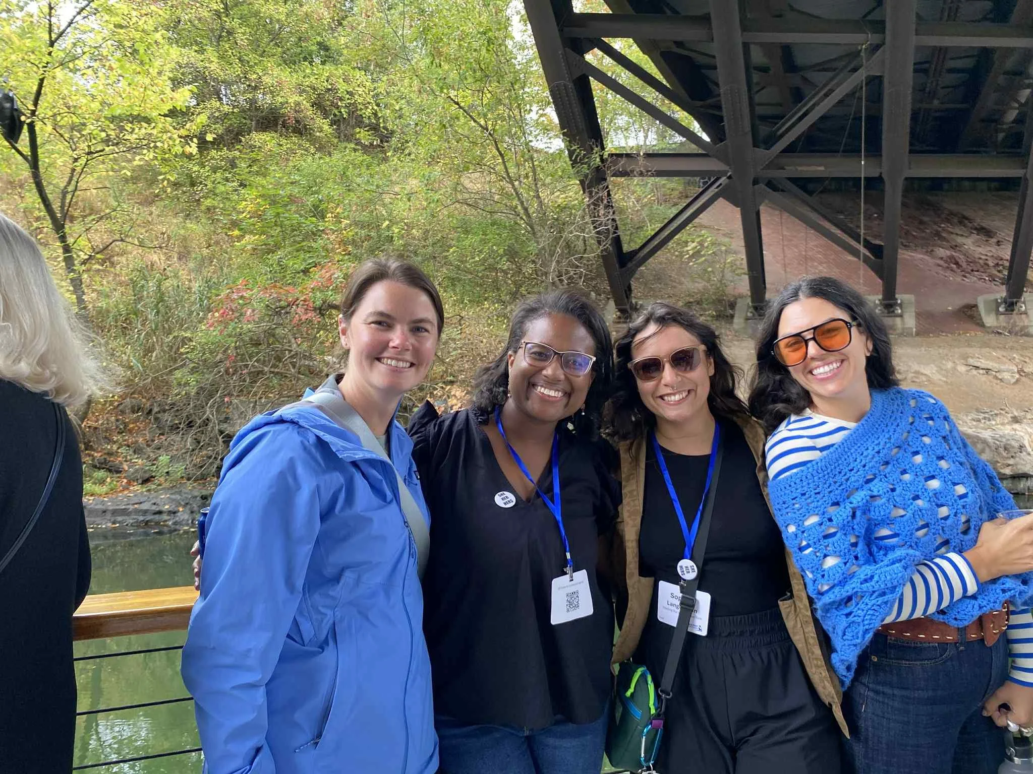 alt="four young women stand together smiling on a boat on a sunny day as it goes under an overpass."