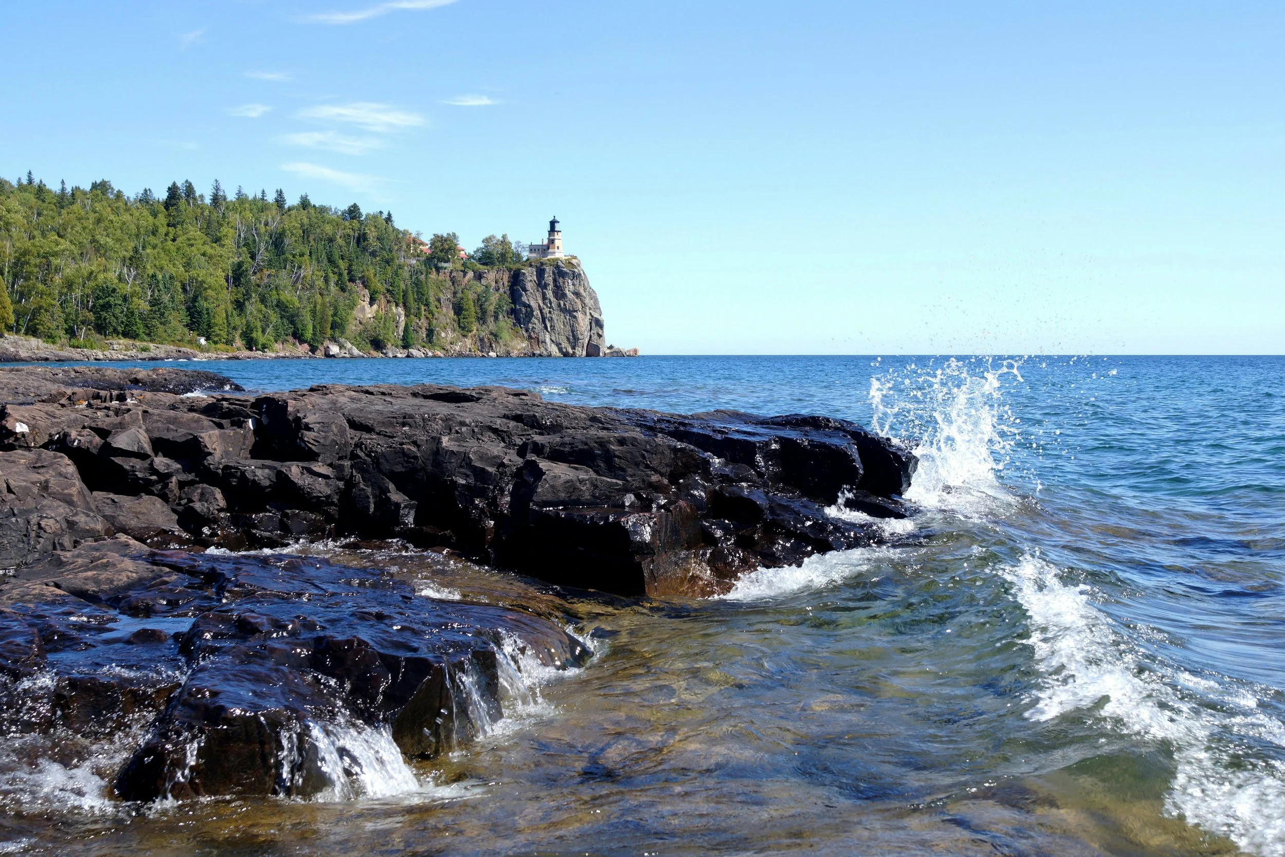 waves crashing against rocks on a lake with green trees and a lighthouse in the background