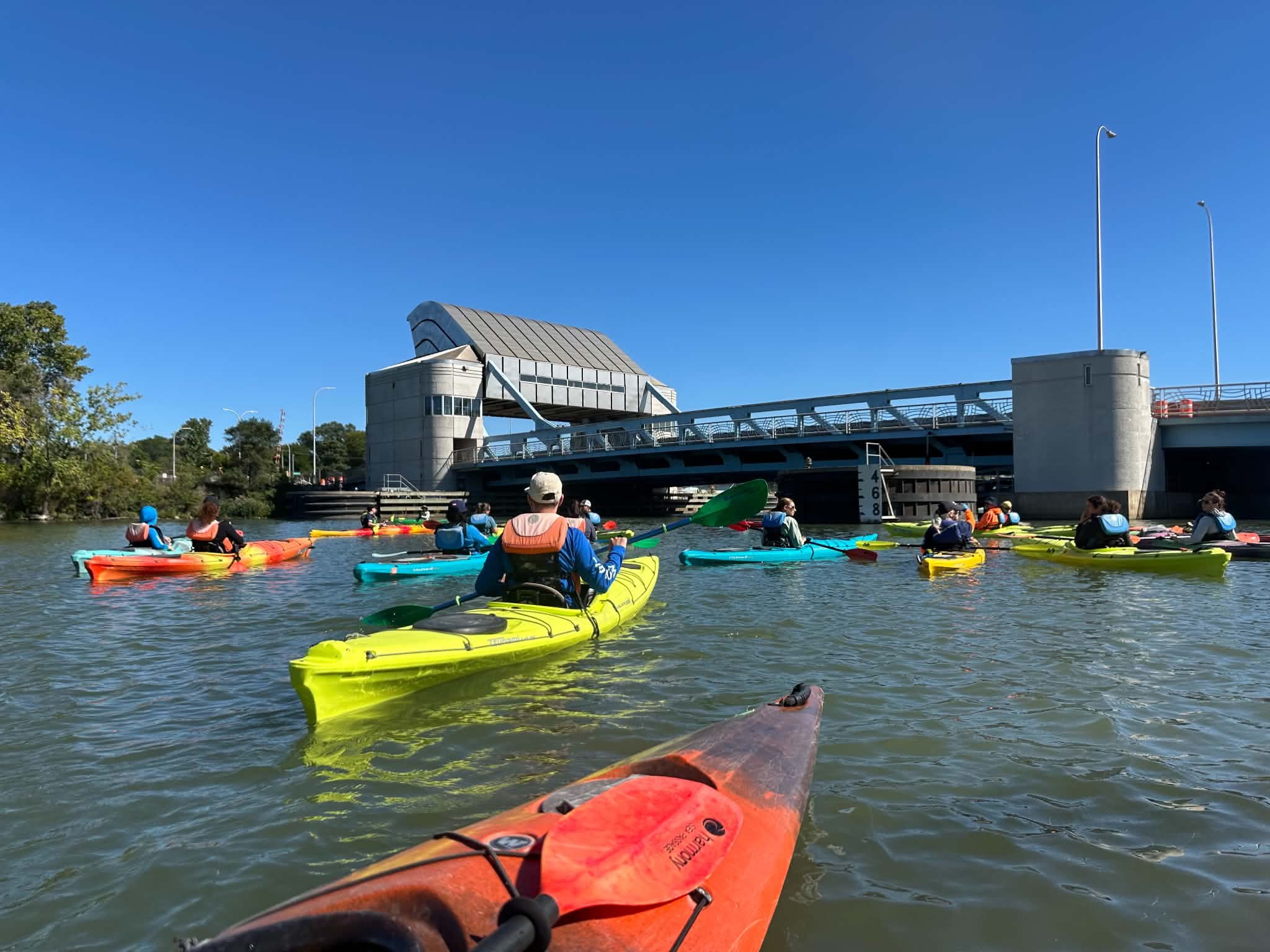 A group of multicolored kayaks prepares to paddle under a bridge.