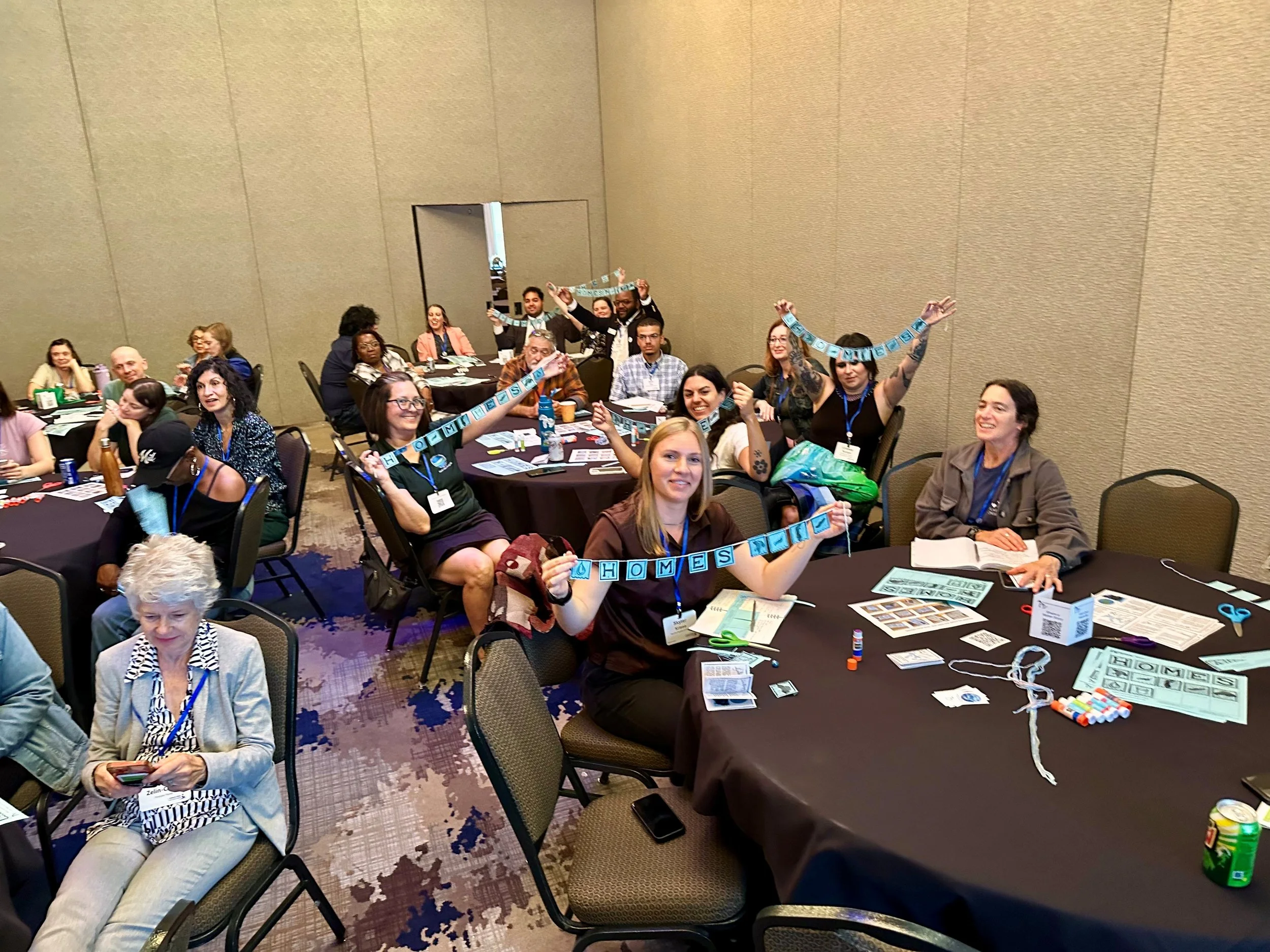 alt="a group of people in a conference room sitting at round tables hold up handmade paper banners that say HOMES to represent the names of the five Great Lakes."